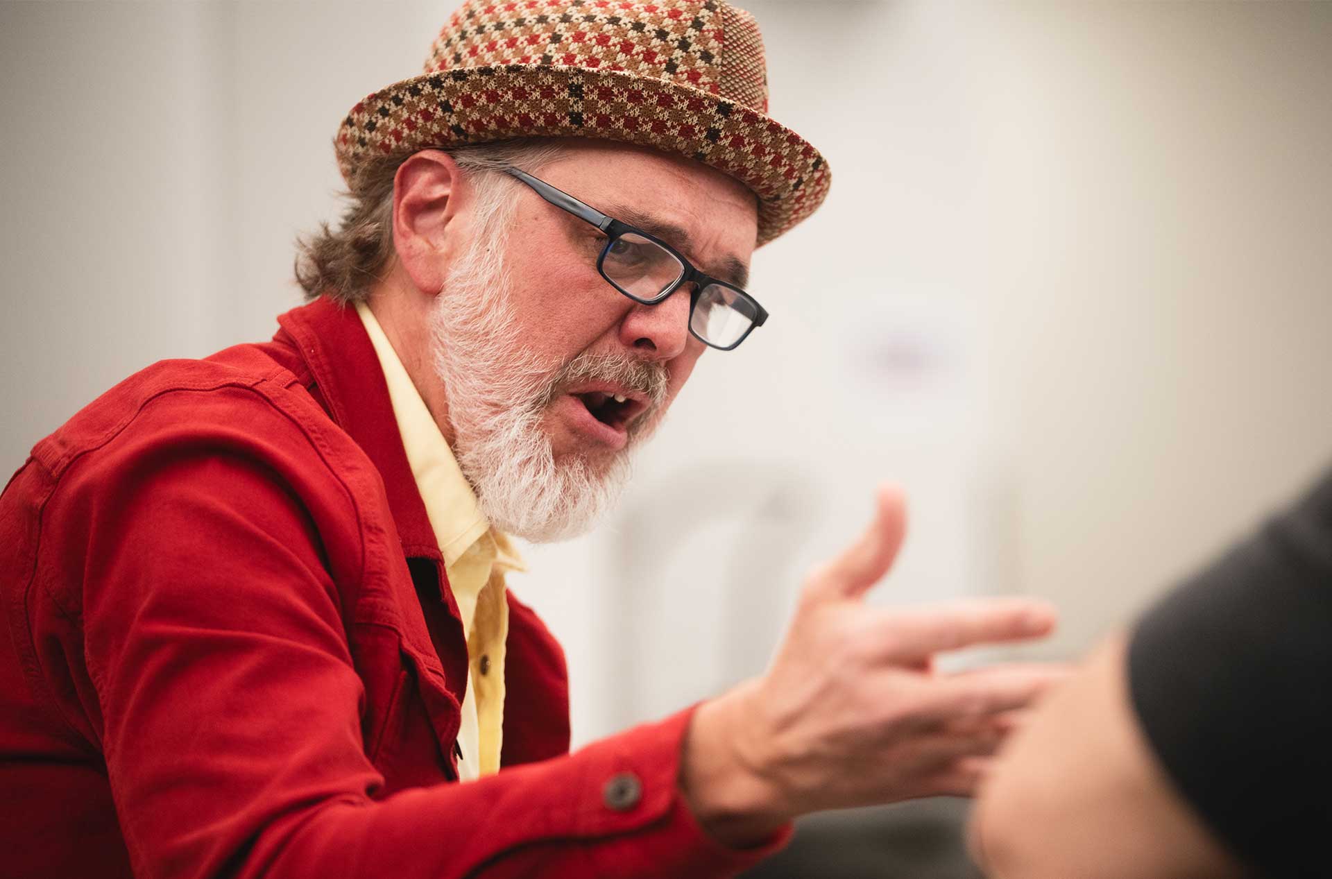 Close-up of a rehearsal participant in a red jacket and patterned hat, speaking passionately and gesturing with one hand.