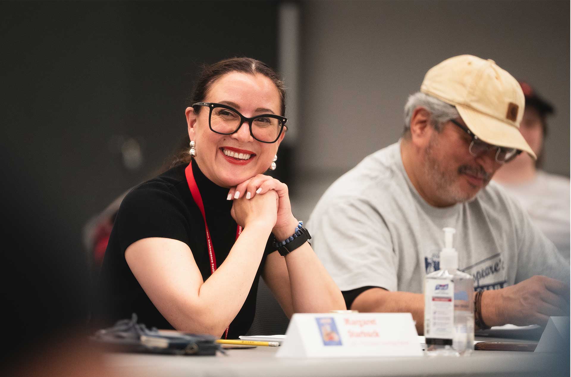 Smiling rehearsal participant sitting at a table, hands resting under the chin, next to a laptop and nameplate; another person beside them types.