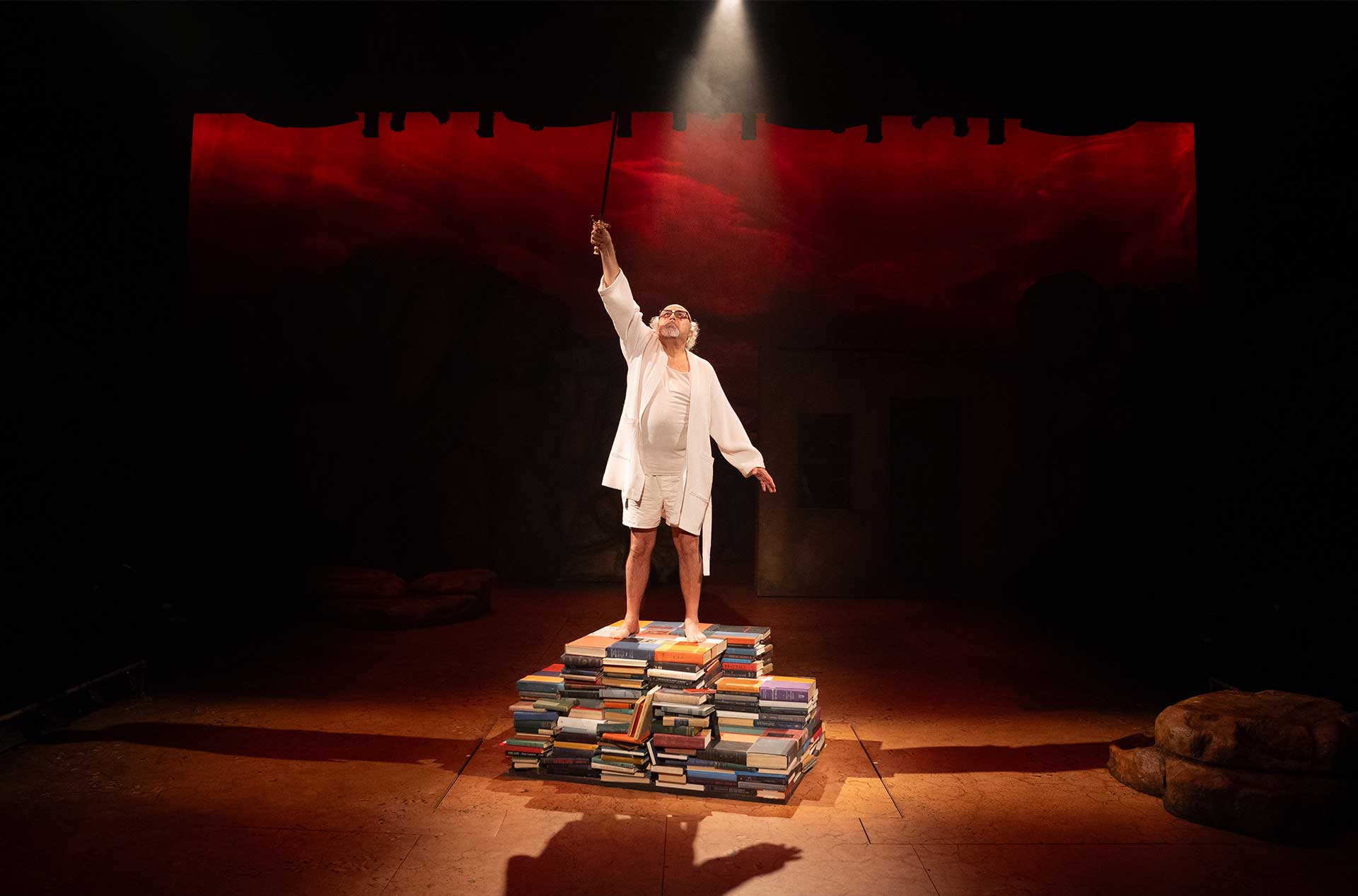 Performer in white stands triumphantly on a stack of books, holding a sword aloft under a spotlight with a dark red sky backdrop.