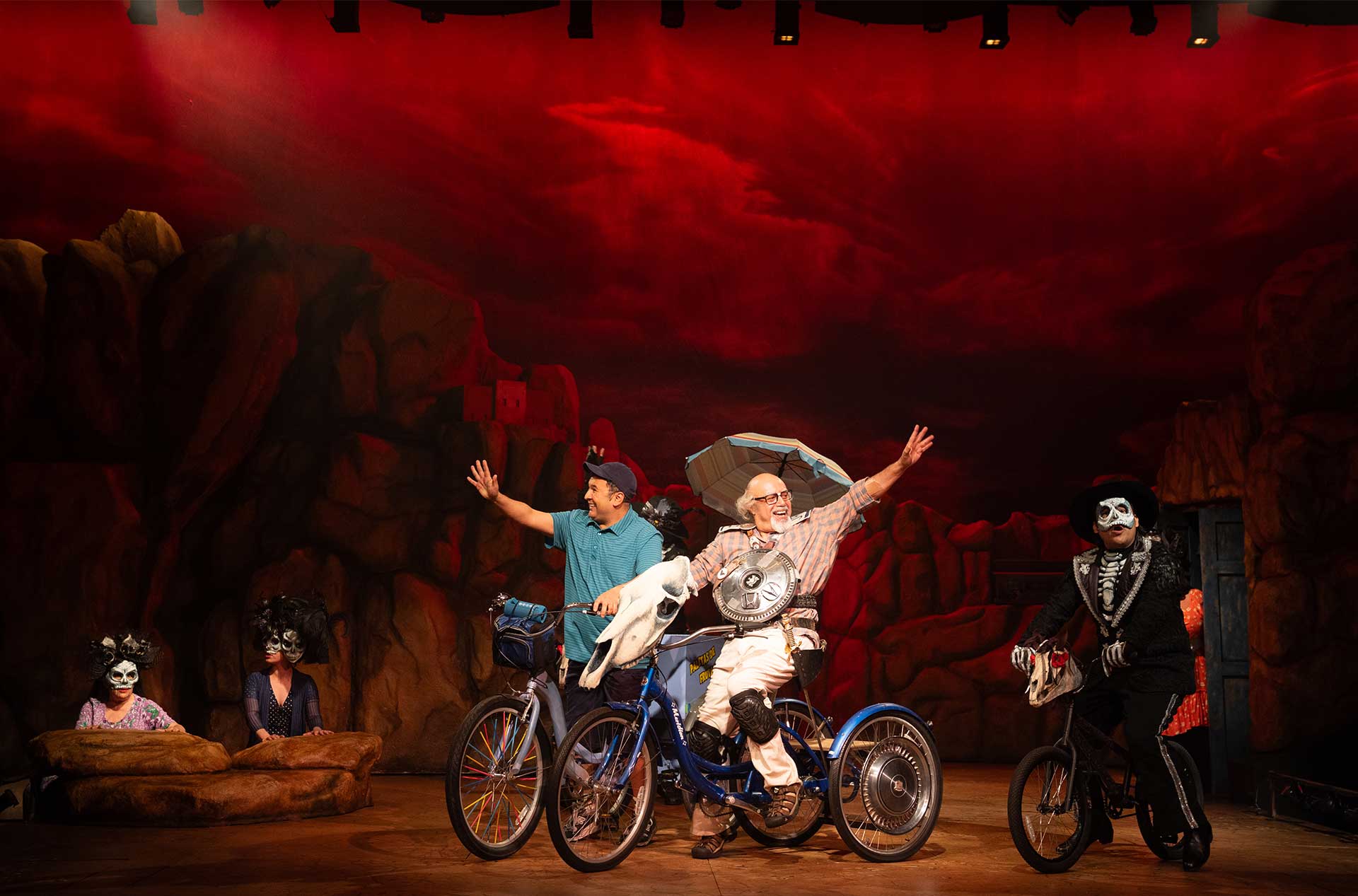 Theatrical scene with characters in skeleton costumes and others riding decorated bicycles against a dramatic red sky backdrop.