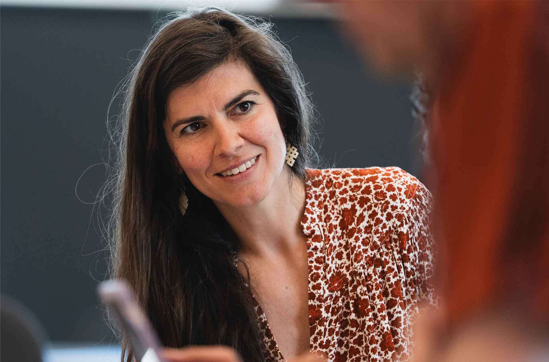 A person with long hair smiles and listens attentively in a close-up shot during a group setting, wearing a red and white patterned top.