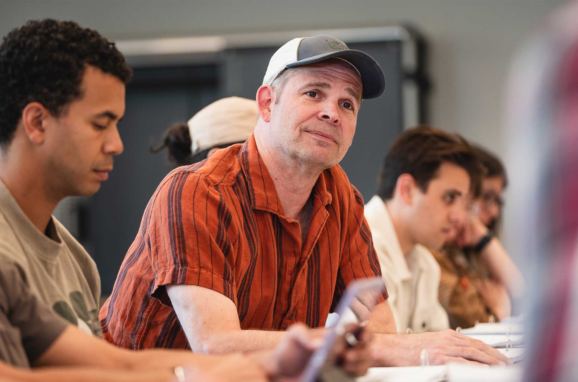 A person in a striped orange shirt and baseball cap sits at a table and listens thoughtfully, surrounded by others focused on their materials.
