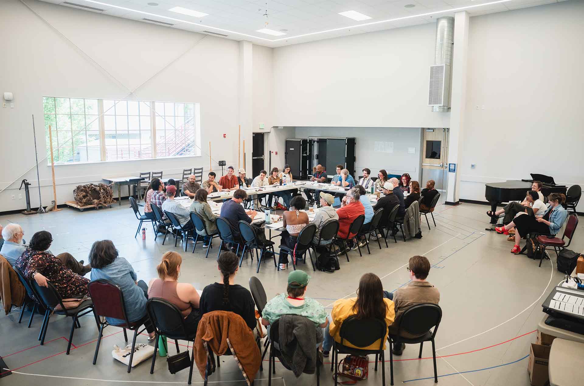 A large group of people sits in a circle of chairs around several connected tables in a spacious room with high ceilings and natural light, engaged in a reheasal.