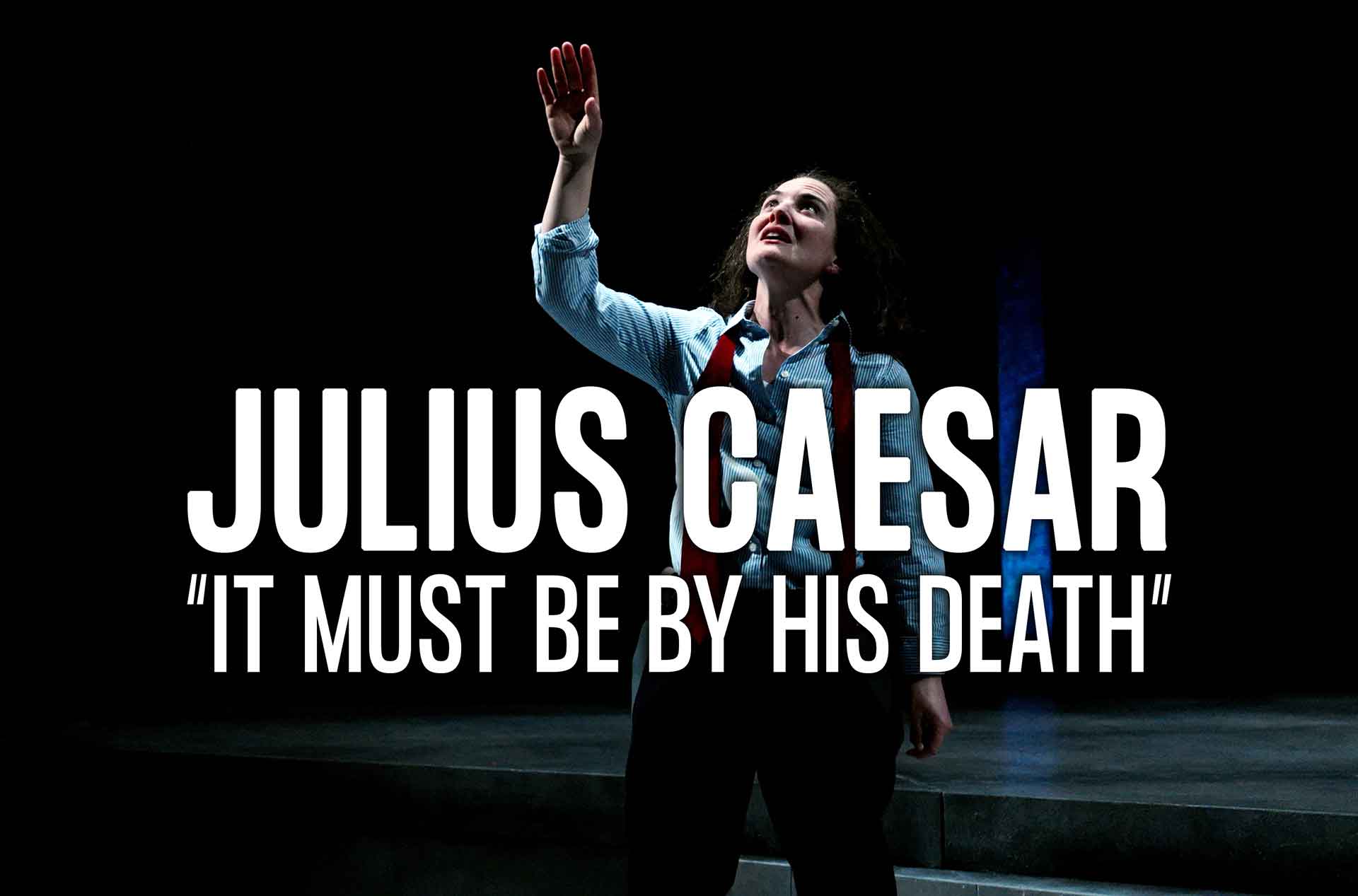 Performer in spotlight on dark stage, wearing light blue shirt, black pants, and red tie, with one arm raised during dramatic monologue from Shakespeare’s Julius Caesar