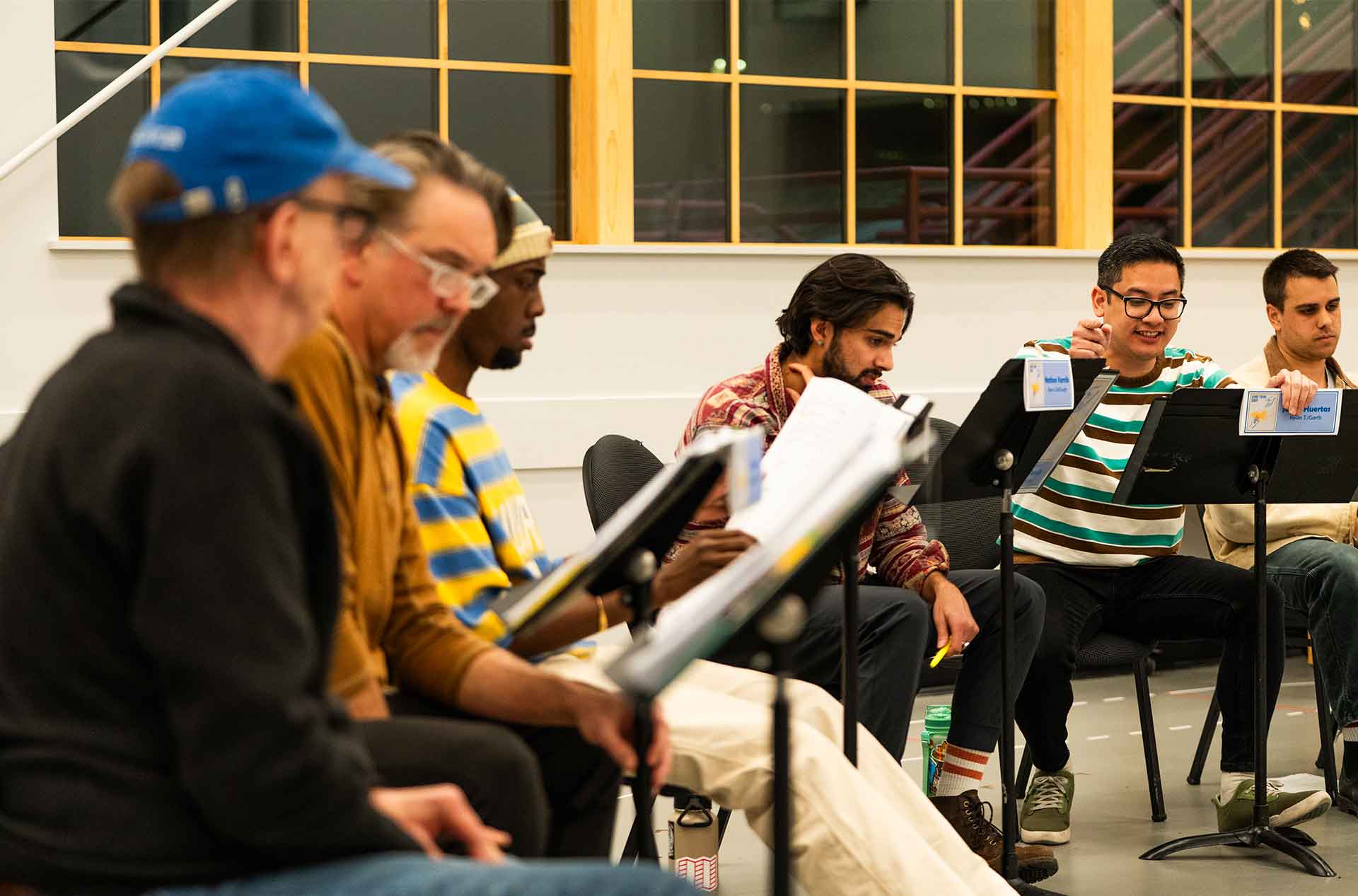 Several participants sit in a row reading from music stands during a rehearsal. Papers, pencils, and focused expressions suggest active review or practice.