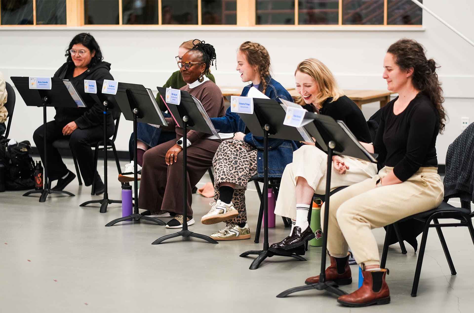 Several participants sit in a line reading from music stands and reacting with smiles and laughter. Water bottles and personal items rest on the floor beneath the stands.