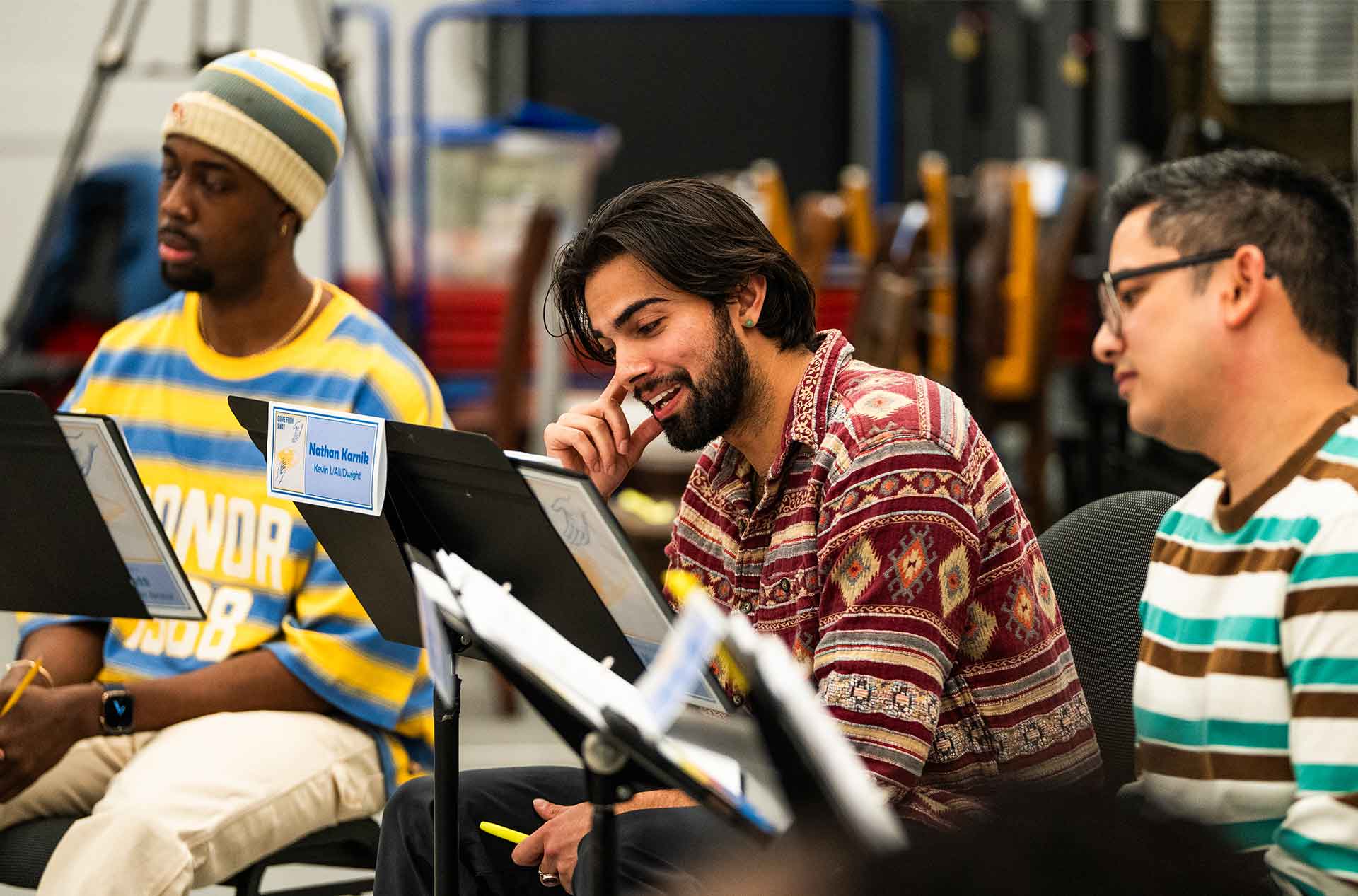 A closer view of participants seated with music stands shows one person gesturing while others read and listen. The setting appears informal and collaborative inside a rehearsal room.