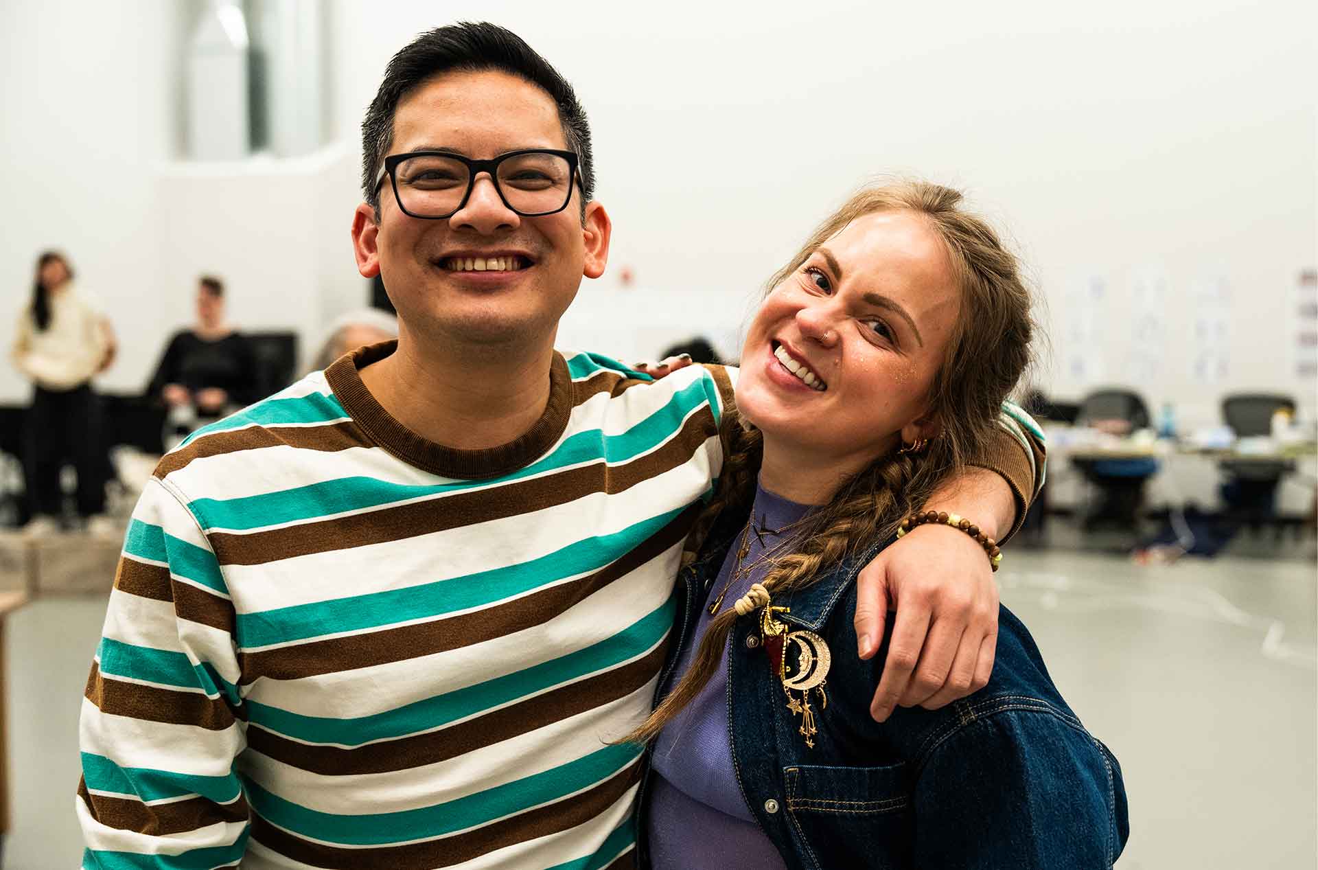 Two people stand close together smiling with arms around each other inside a large rehearsal space. Music stands and other participants are visible blurred in the background, suggesting a collaborative setting.