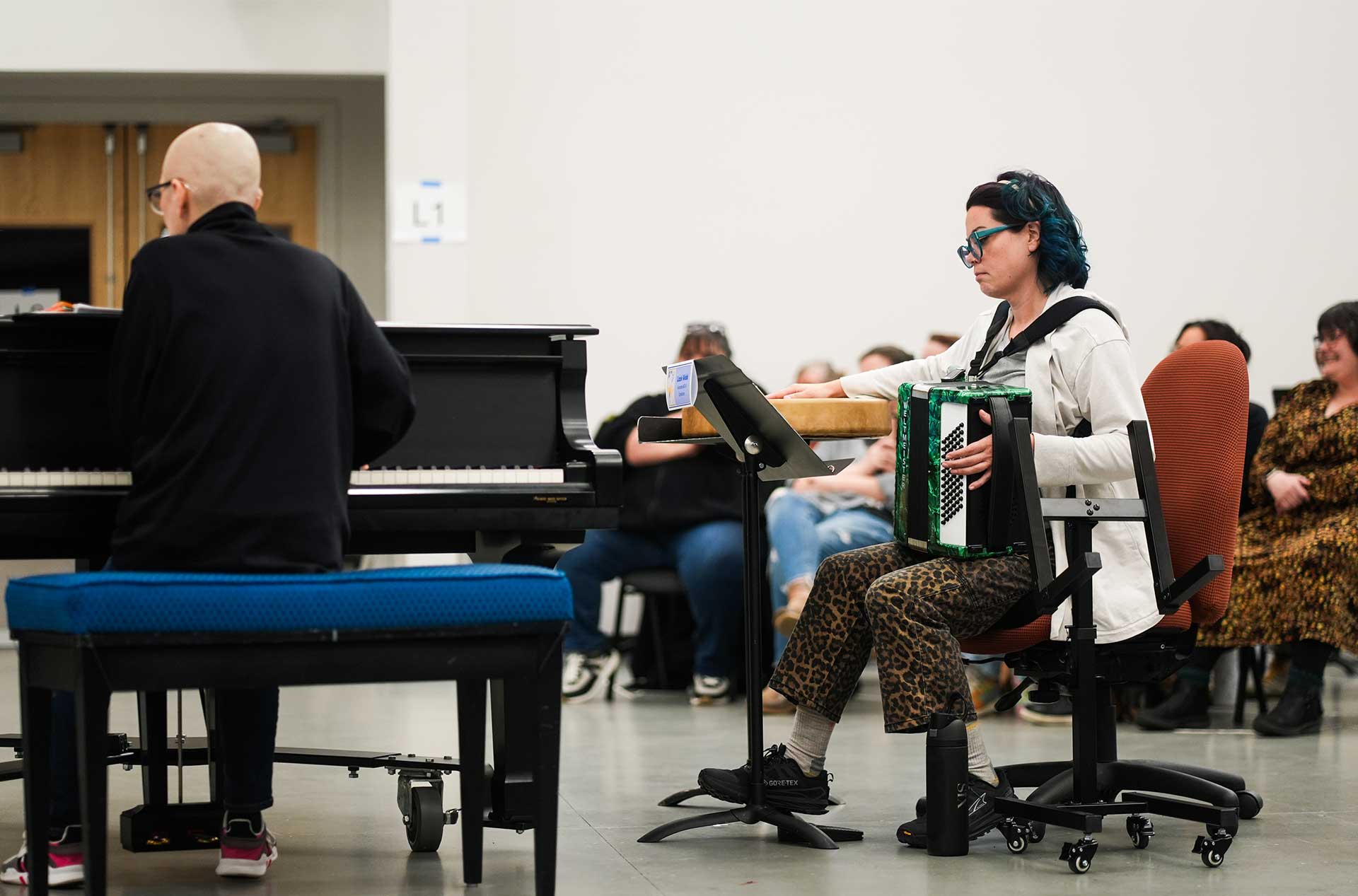 One person plays a grand piano while another sits nearby holding an accordion, both focused on the music. Other participants sit in the background observing the rehearsal.