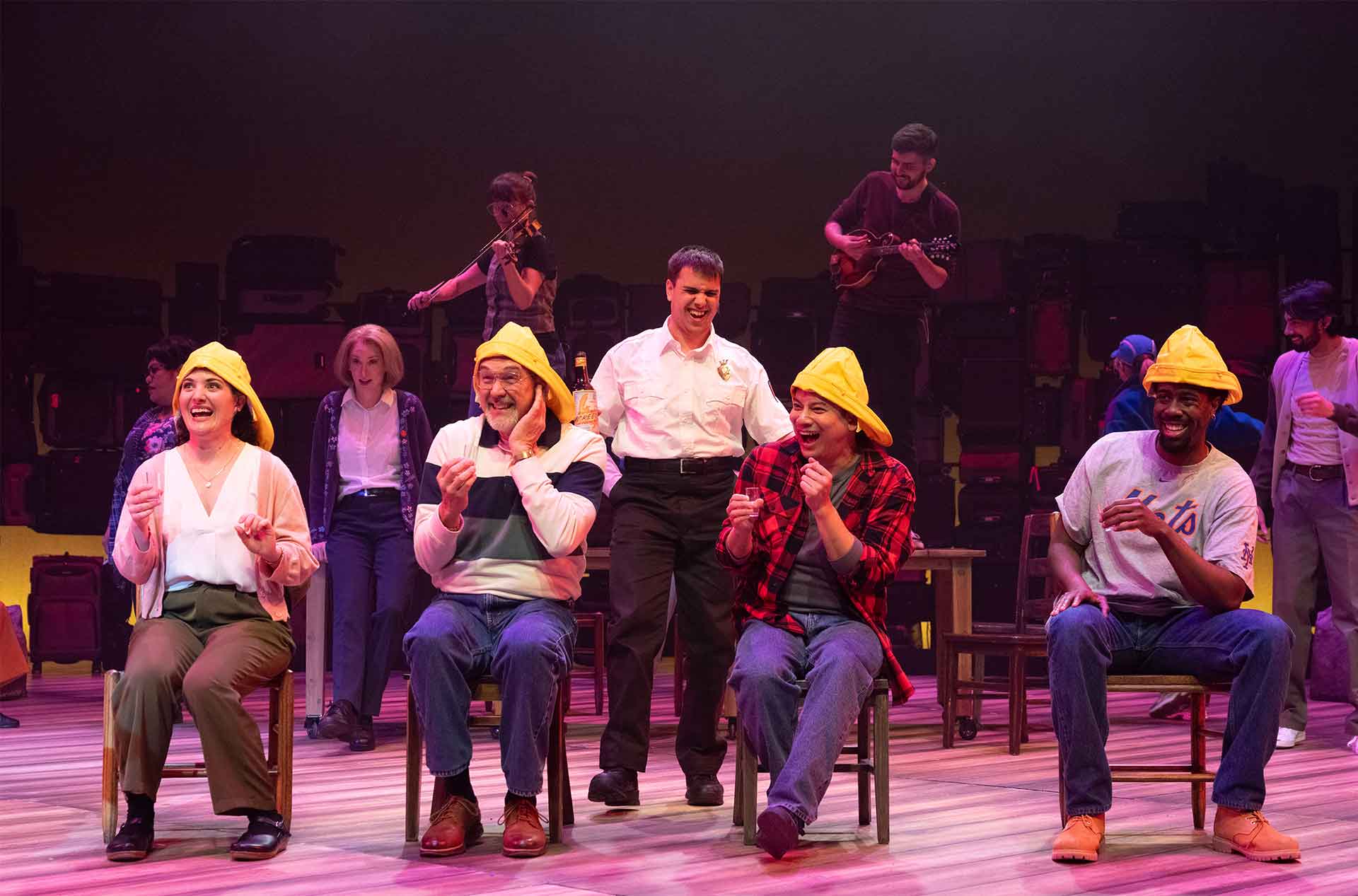 Several seated performers laugh and react together while musicians play behind them on stage.