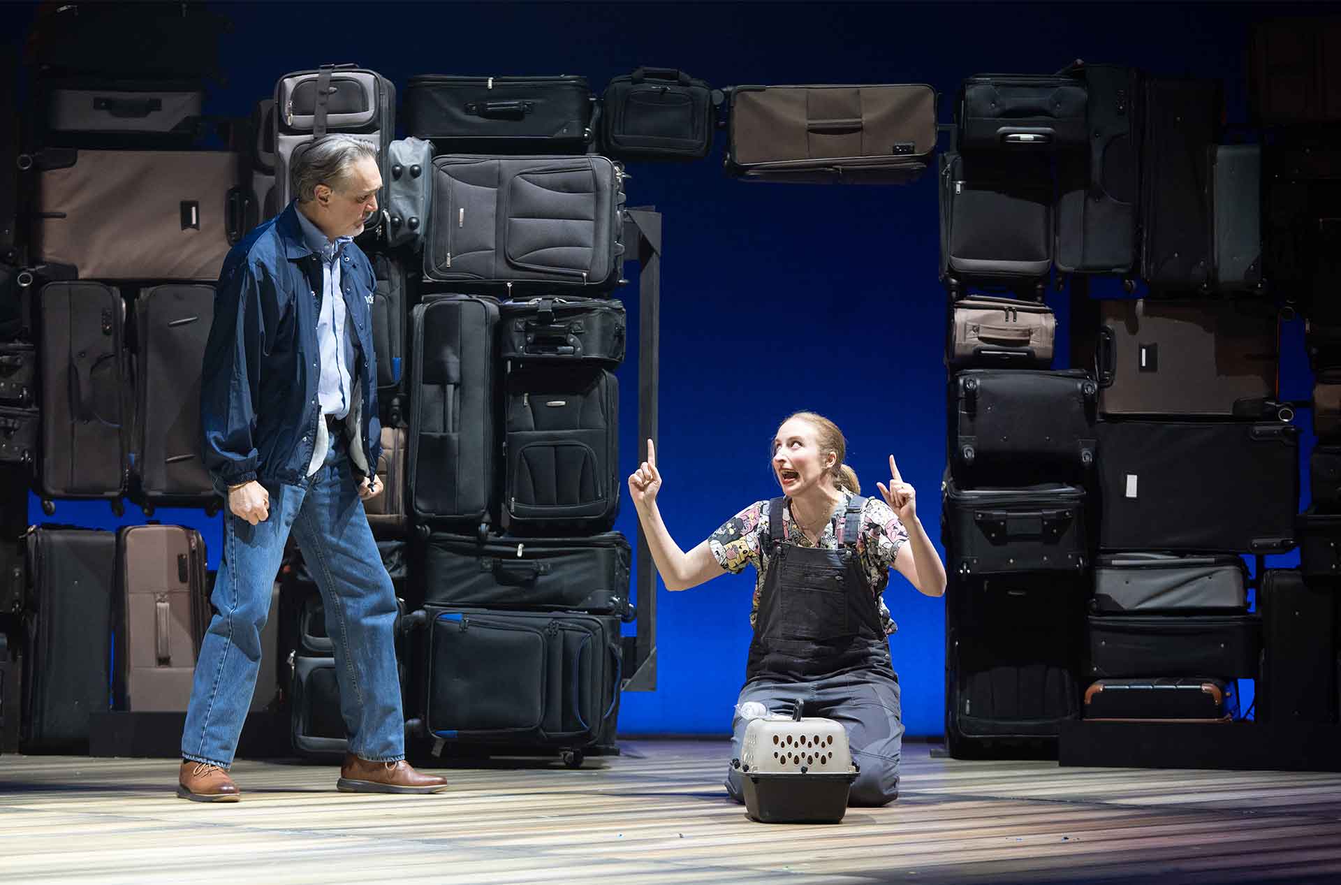 One person kneels on stage pointing upward beside a pet carrier while another stands nearby, with stacked suitcases forming the backdrop.