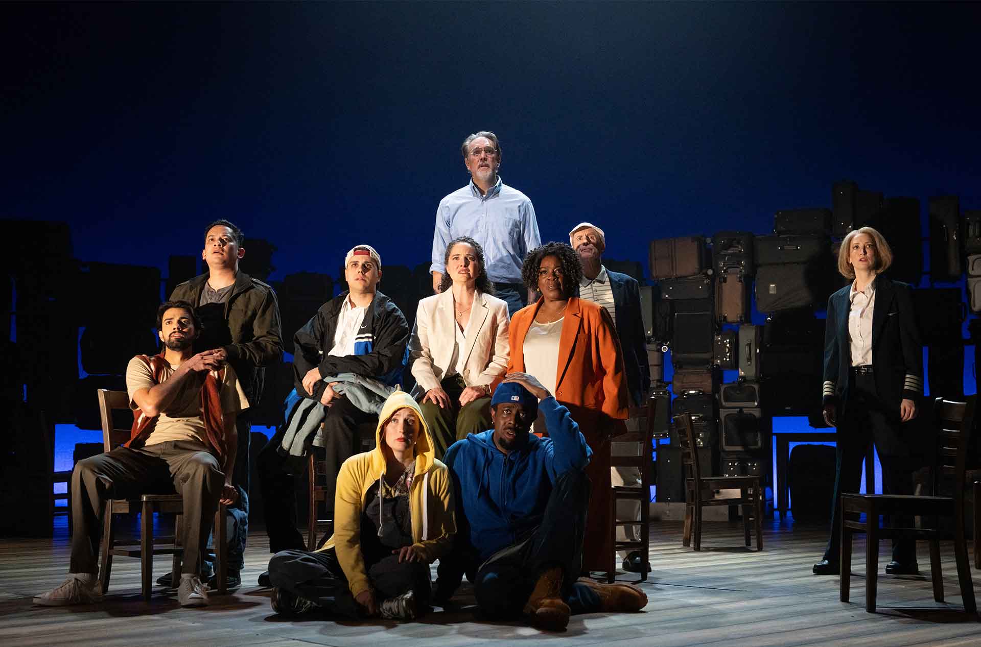 A group of people sit and stand together on stage looking forward, framed by rows of stacked luggage.