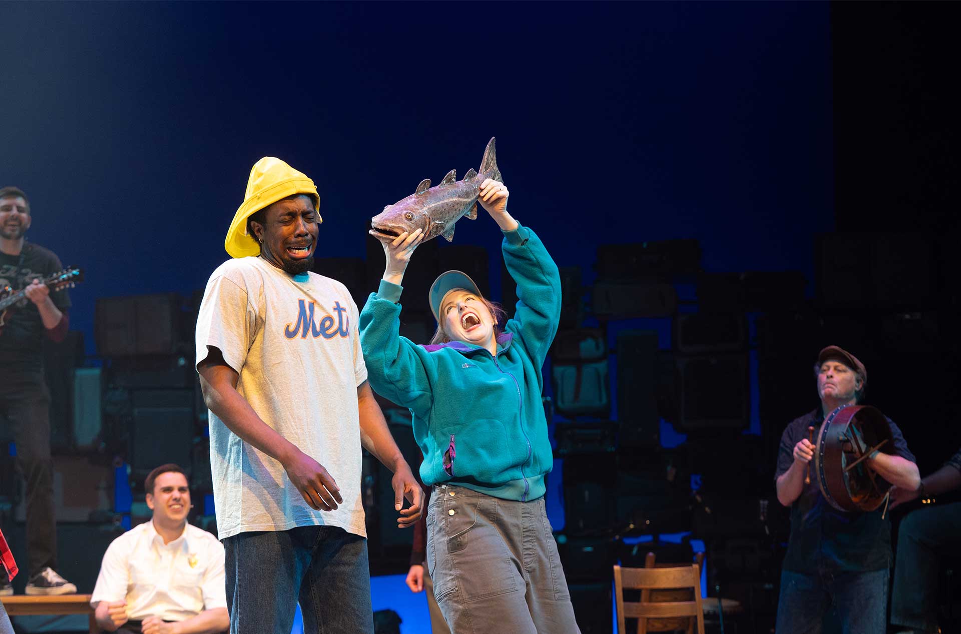 One person lifts a fish overhead while another reacts beside them, with musicians visible in the background.
