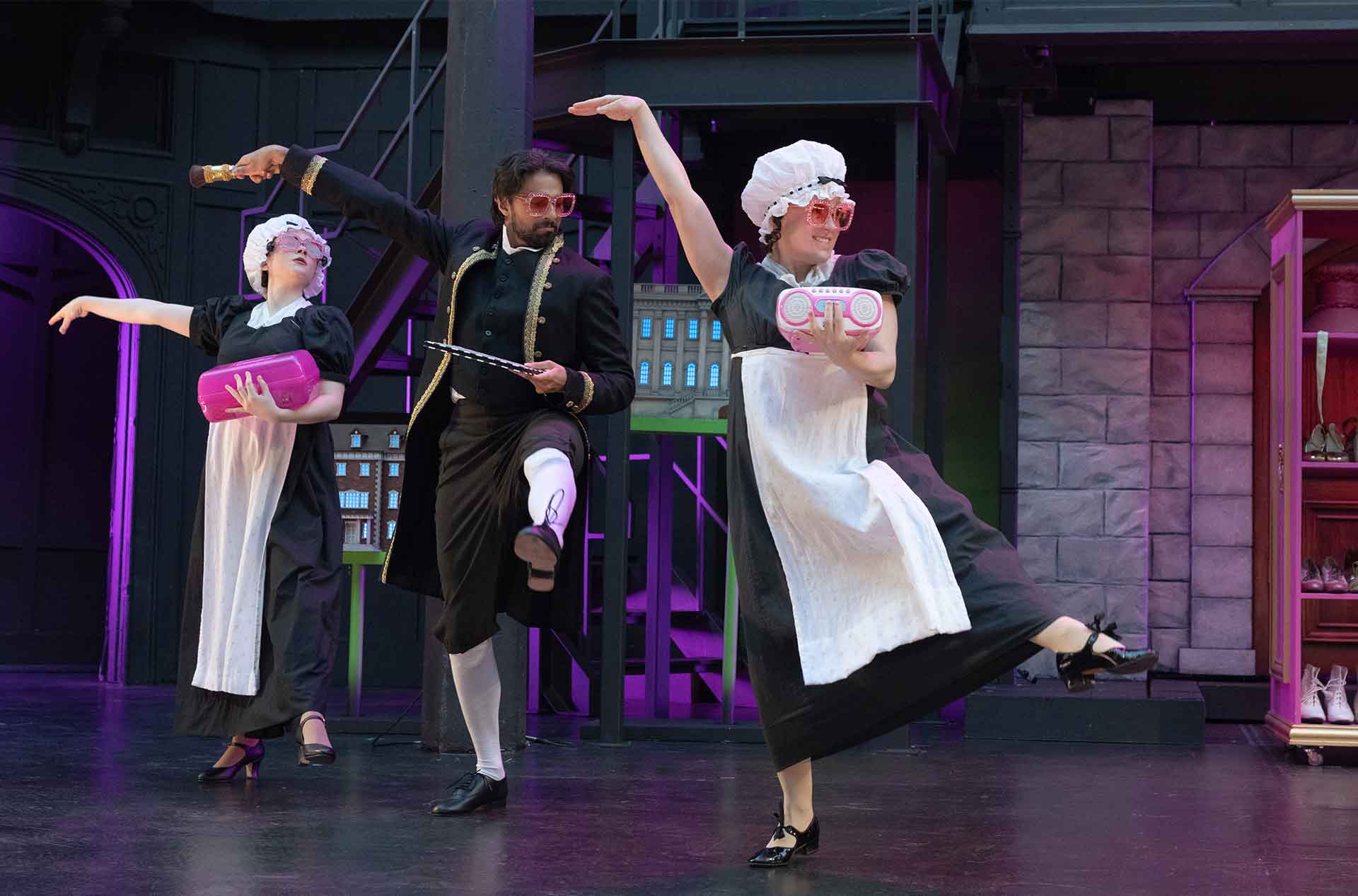 Three performers dance in playful maid and formal costumes with oversized pink glasses. Two carry bright pink props while the center performer holds a tray and small gold object.