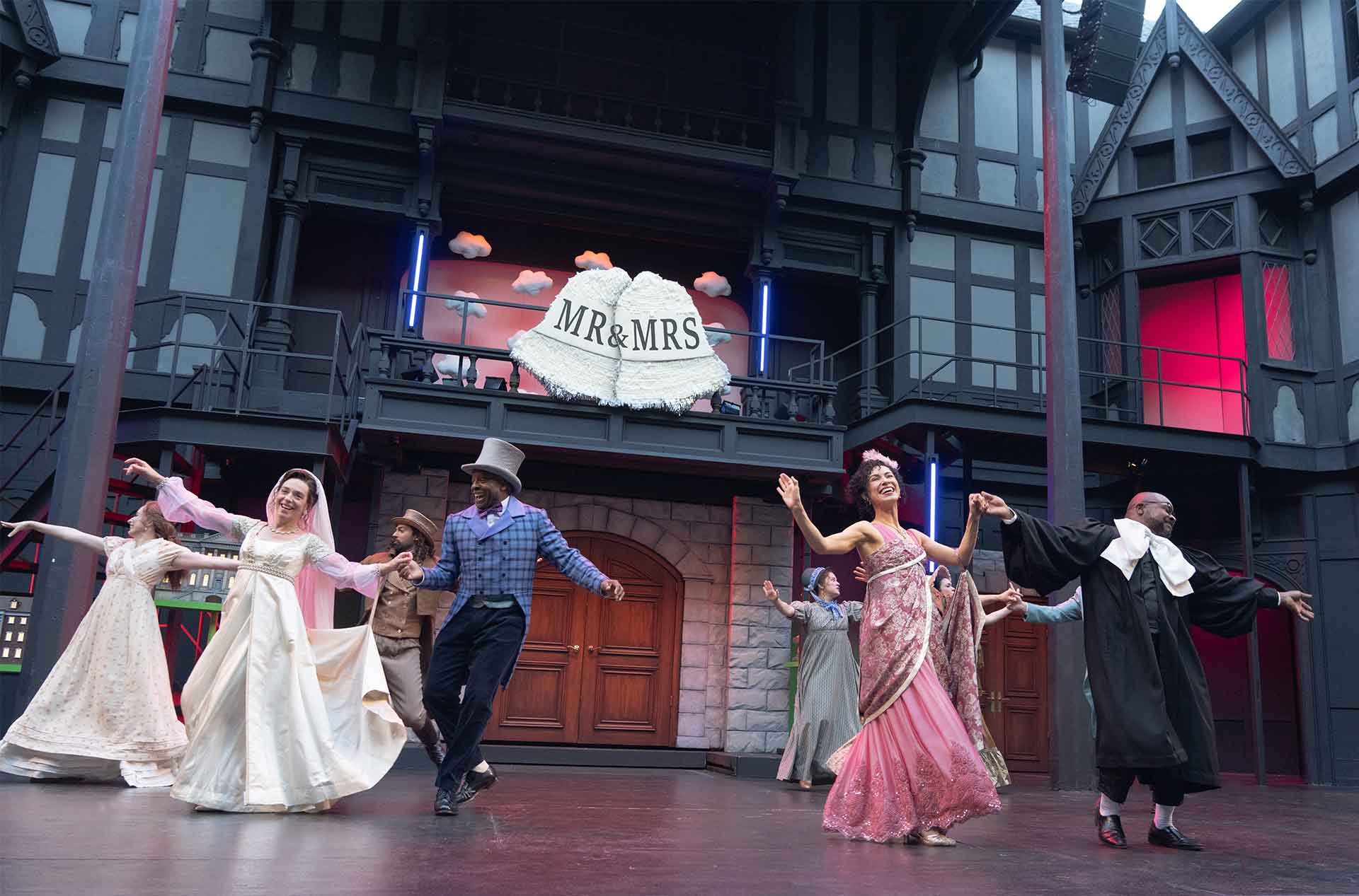 A theater ensemble dances in wedding attire beneath a large bell that reads "MR&MRS". The stage set includes gray house facades, balconies, and glowing red windows.