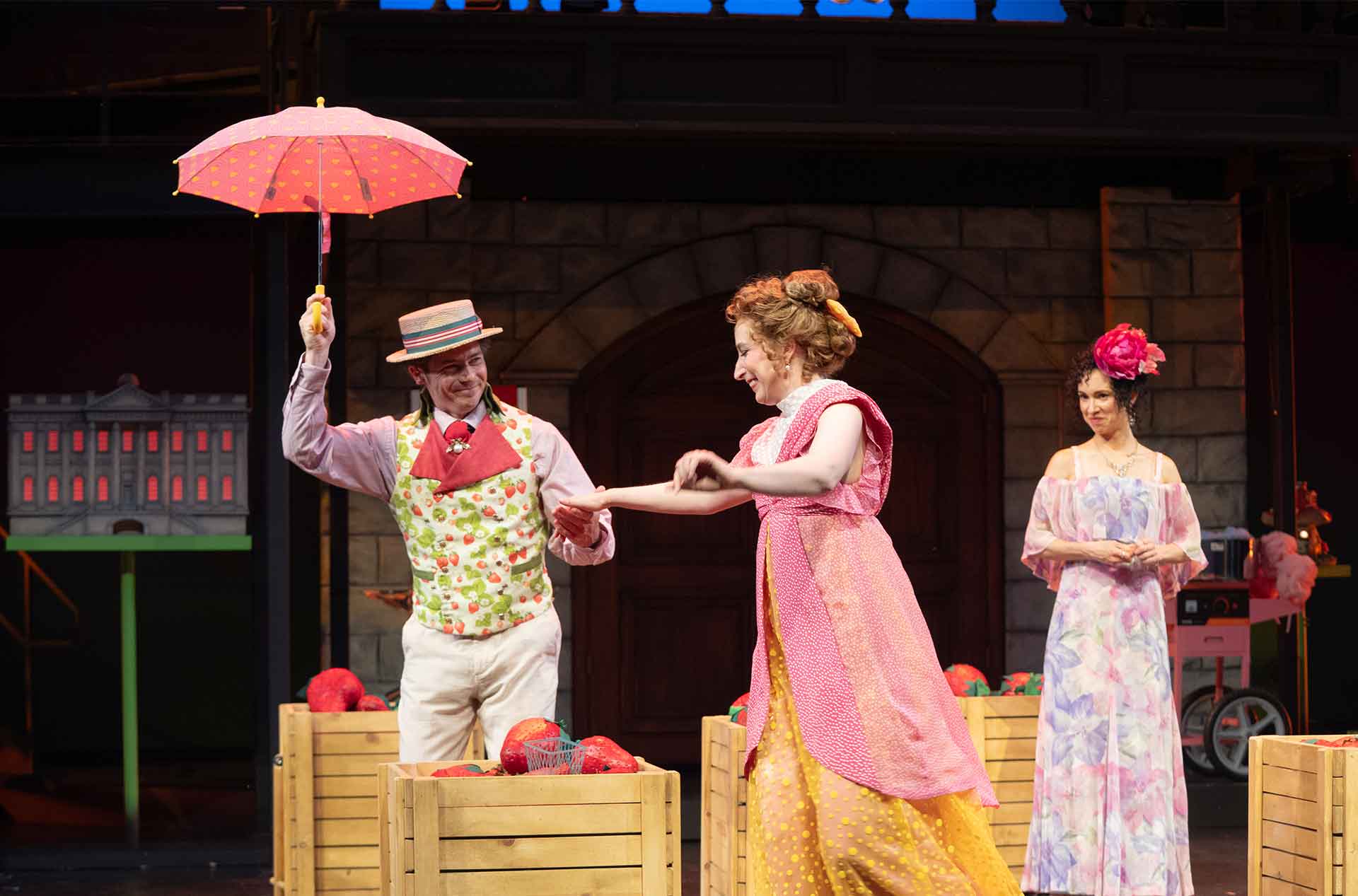 Three performers stand among crates of red props on a theater stage. One holds hands with another performer while a third watches from behind.