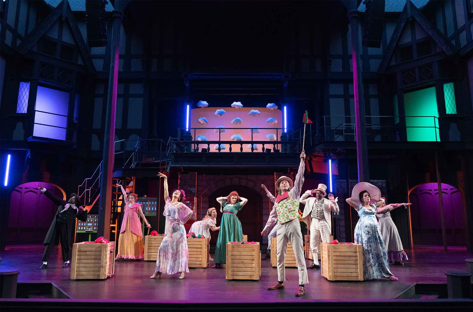 A theater ensemble performs across a bright stage with wooden crates and colorful period costumes. The set includes tall house facades, glowing panels, and a balcony with cloud props.