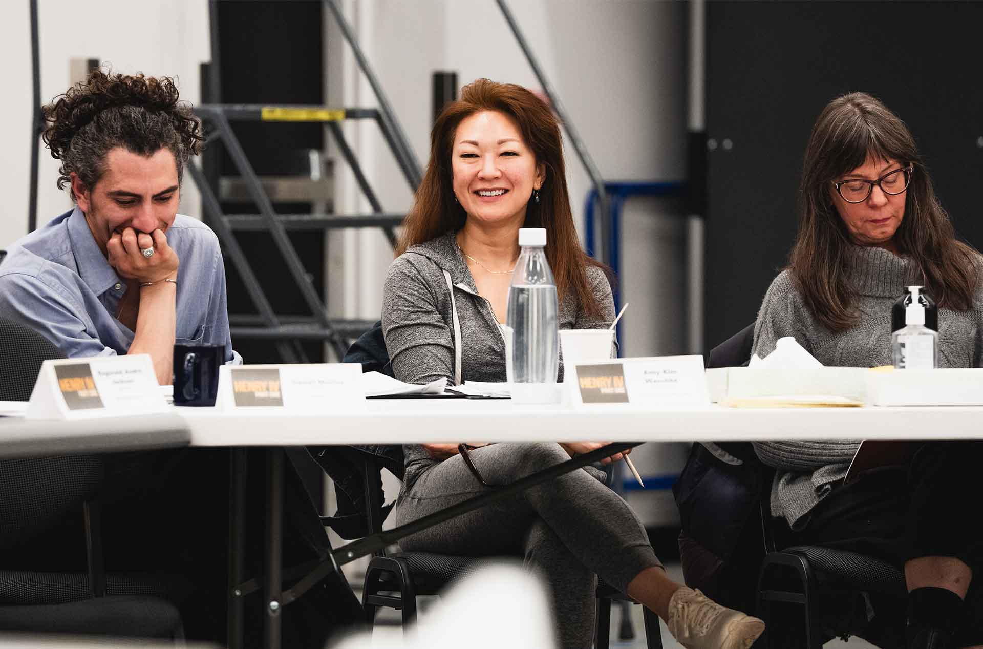 Three cast members seated at a rehearsal table smiling during a collaborative play rehearsal.