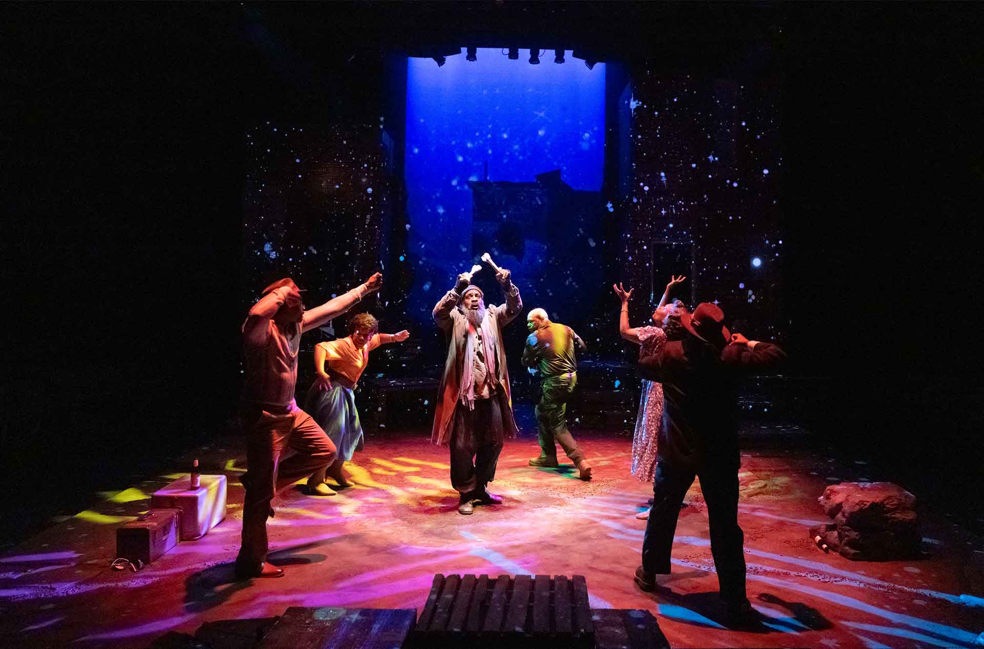 Six performers move with raised arms beneath vivid red and blue stage lighting. A star filled backdrop creates a dreamlike atmosphere around the group.
