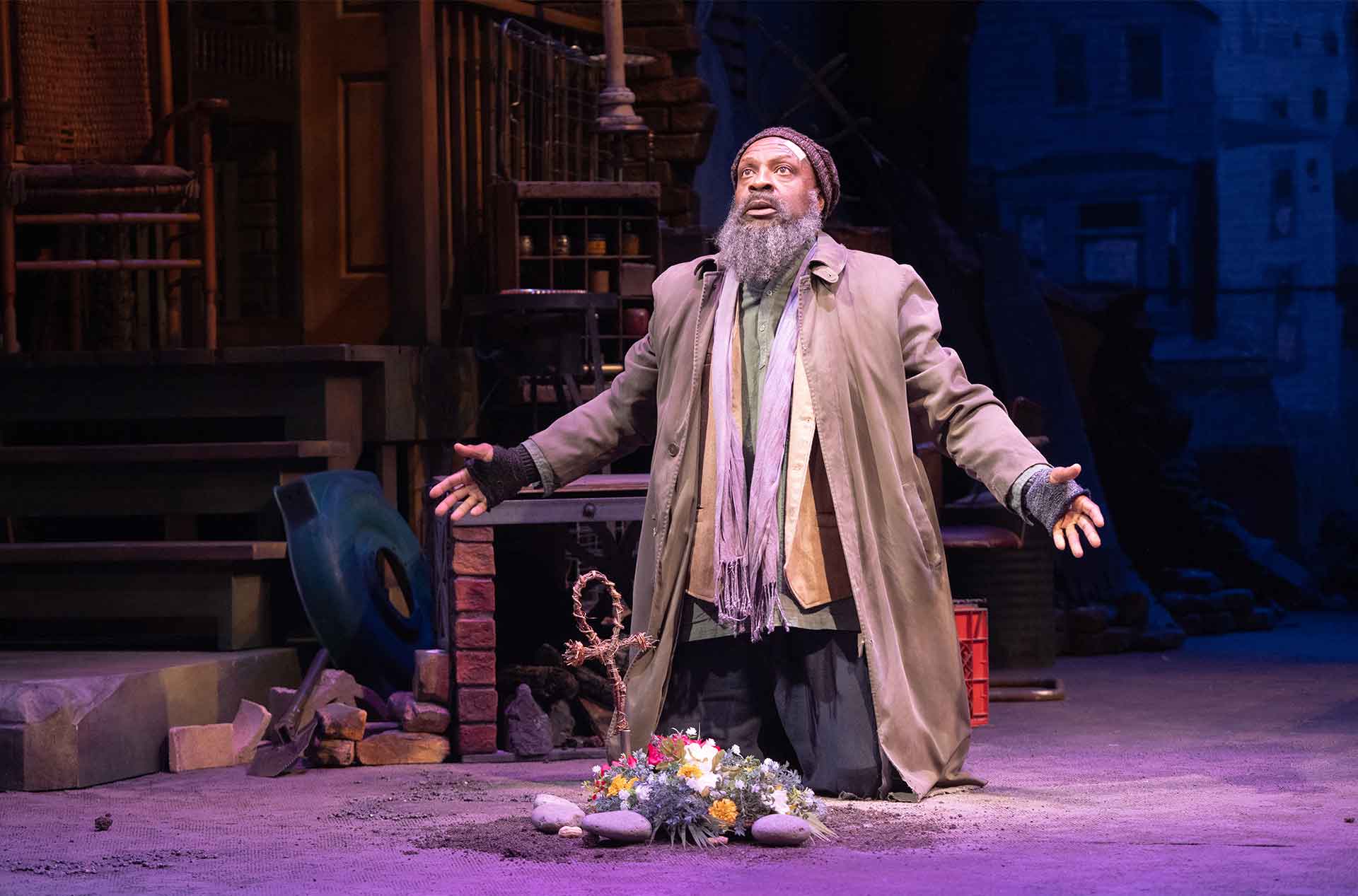 A bearded performer kneels with arms spread beside flowers arranged within a ring of stones. Their upward gaze and open posture create a solemn moment.