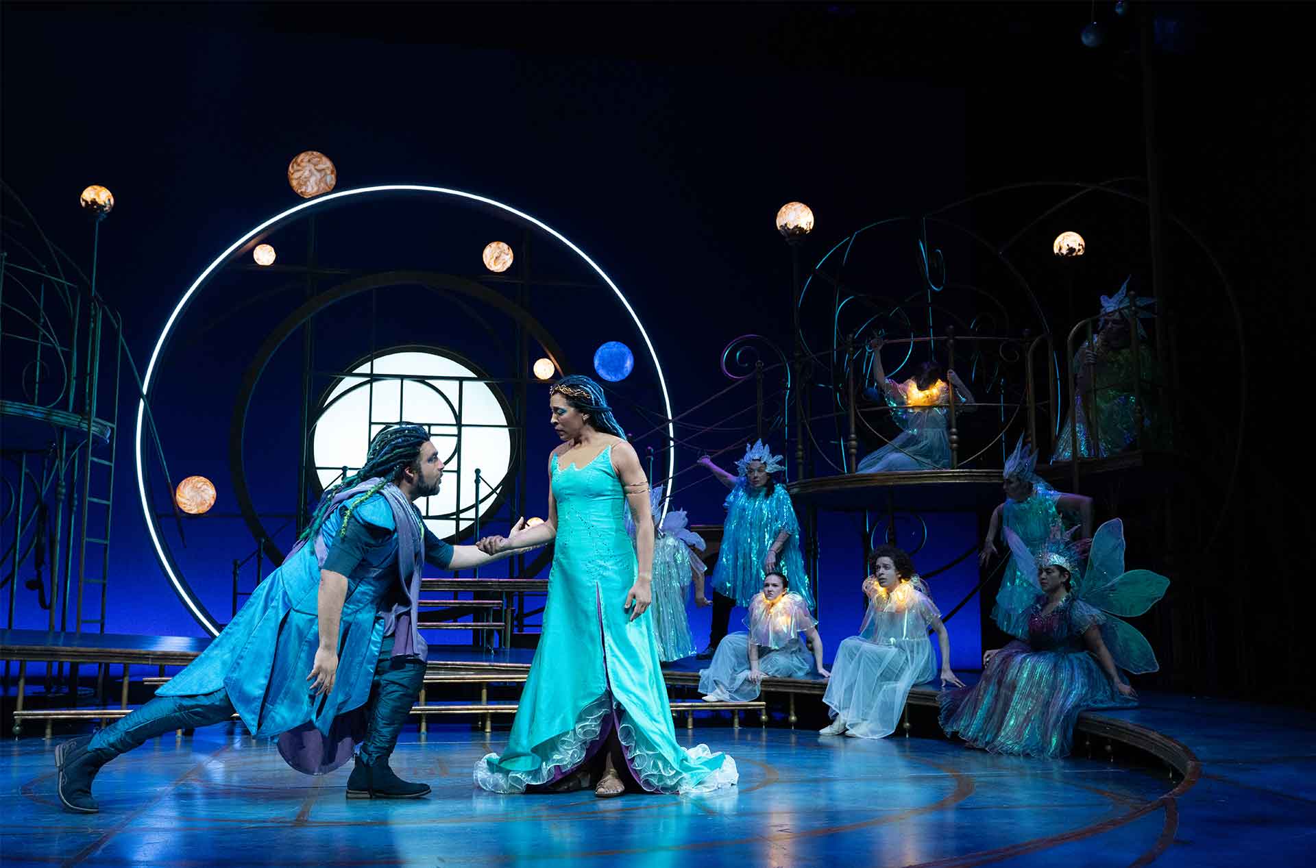Two performers hold hands at center stage in a dramatic exchange while ensemble members in whimsical costumes watch from behind.