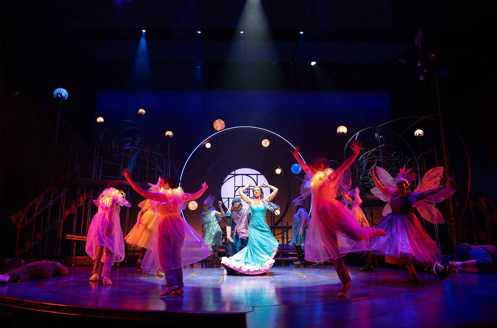 Performer in a flowing blue dress dances center stage surrounded by ensemble cast in colorful costumes under glowing circular lights on a theatrical set.