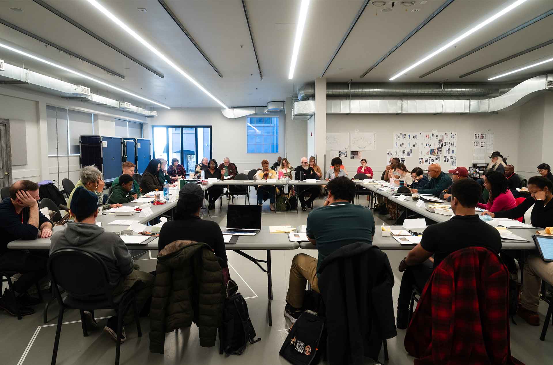 Wide shot of a large group seated around a square table setup for a play read-through in a bright rehearsal room.