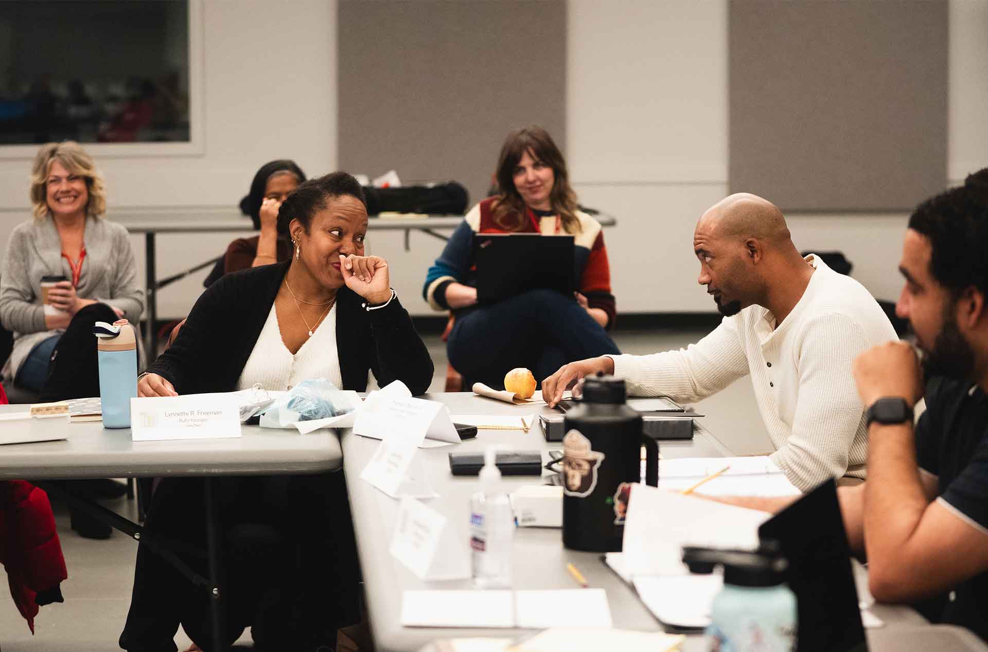 People sitting around a table engaged in lively conversation during a script read-through, with name cards, water bottles, and scripts visible.