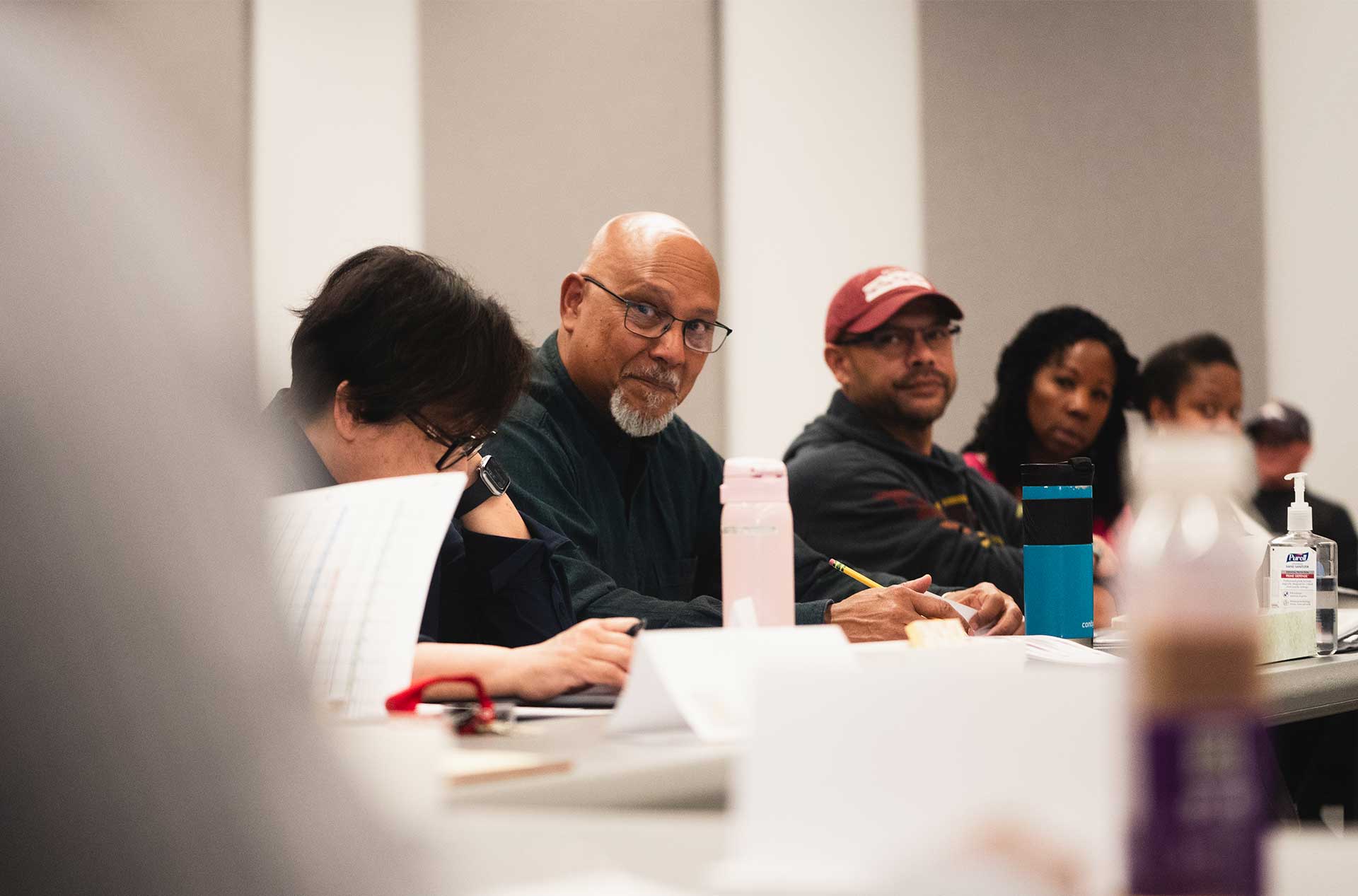 Person looking toward the camera while seated at a table surrounded by others reading and taking notes during rehearsal.