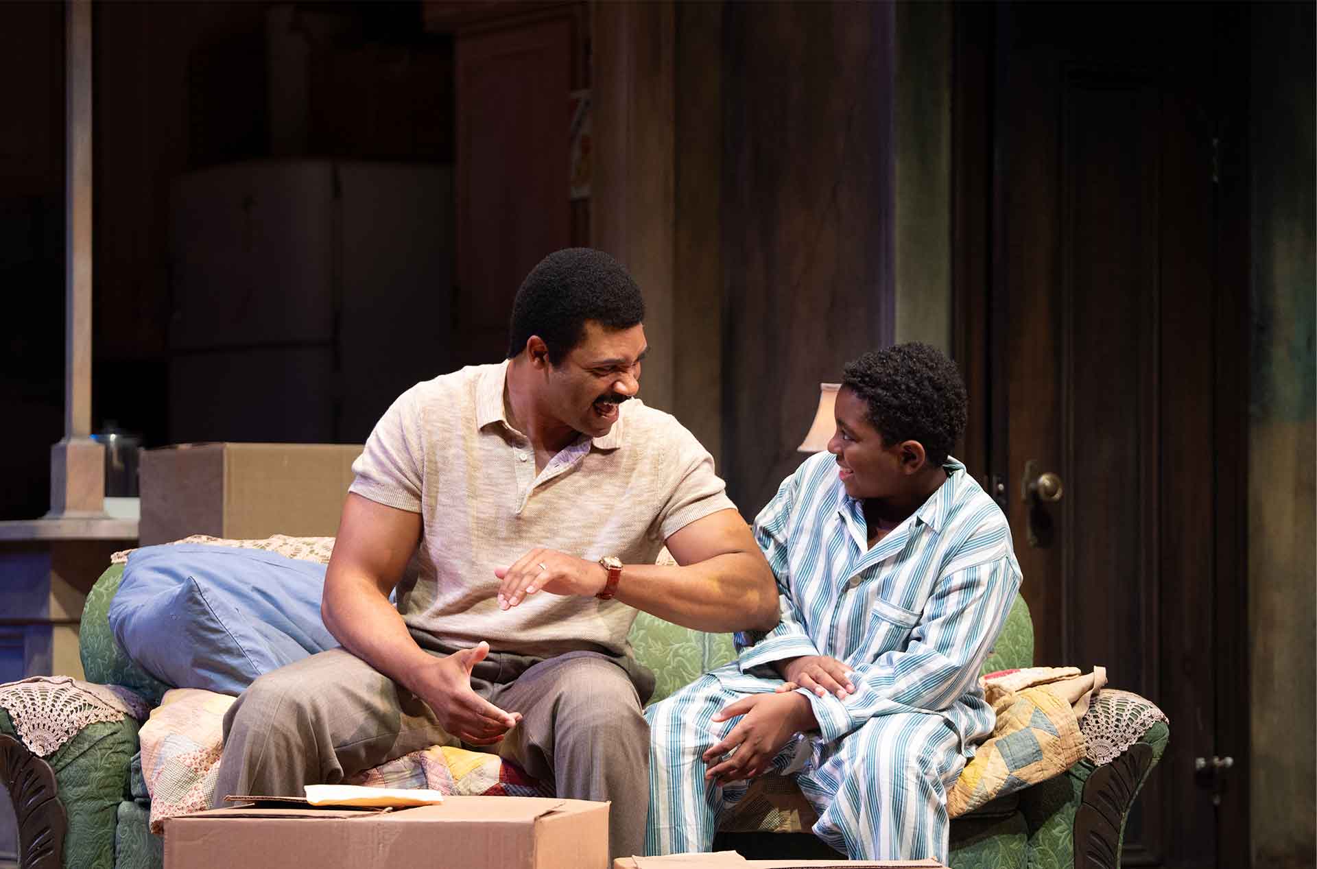 Two people sit on a couch smiling and talking, with moving boxes and a cozy home setting around them.