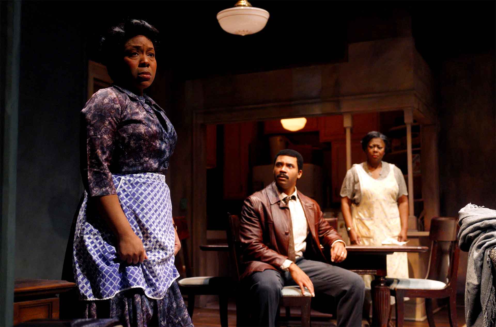 Three people in a dimly lit home interior react tensely during a conversation, with one woman standing in the foreground wearing a patterned dress and apron while a man and another woman stand near a dining table behind her.