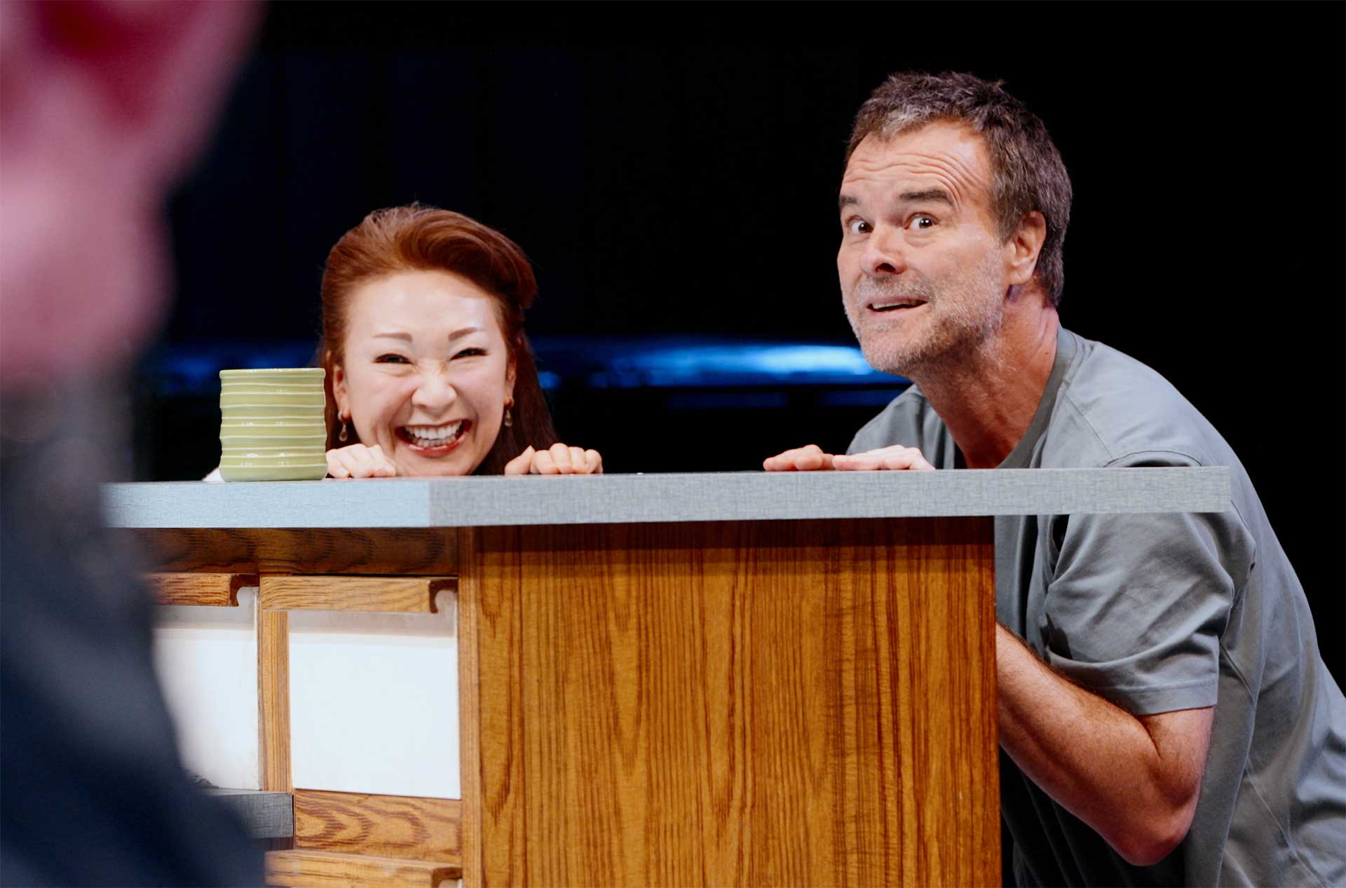 Two performers crouch behind a wooden counter onstage. One laughs while the other looks up with a surprised expression.