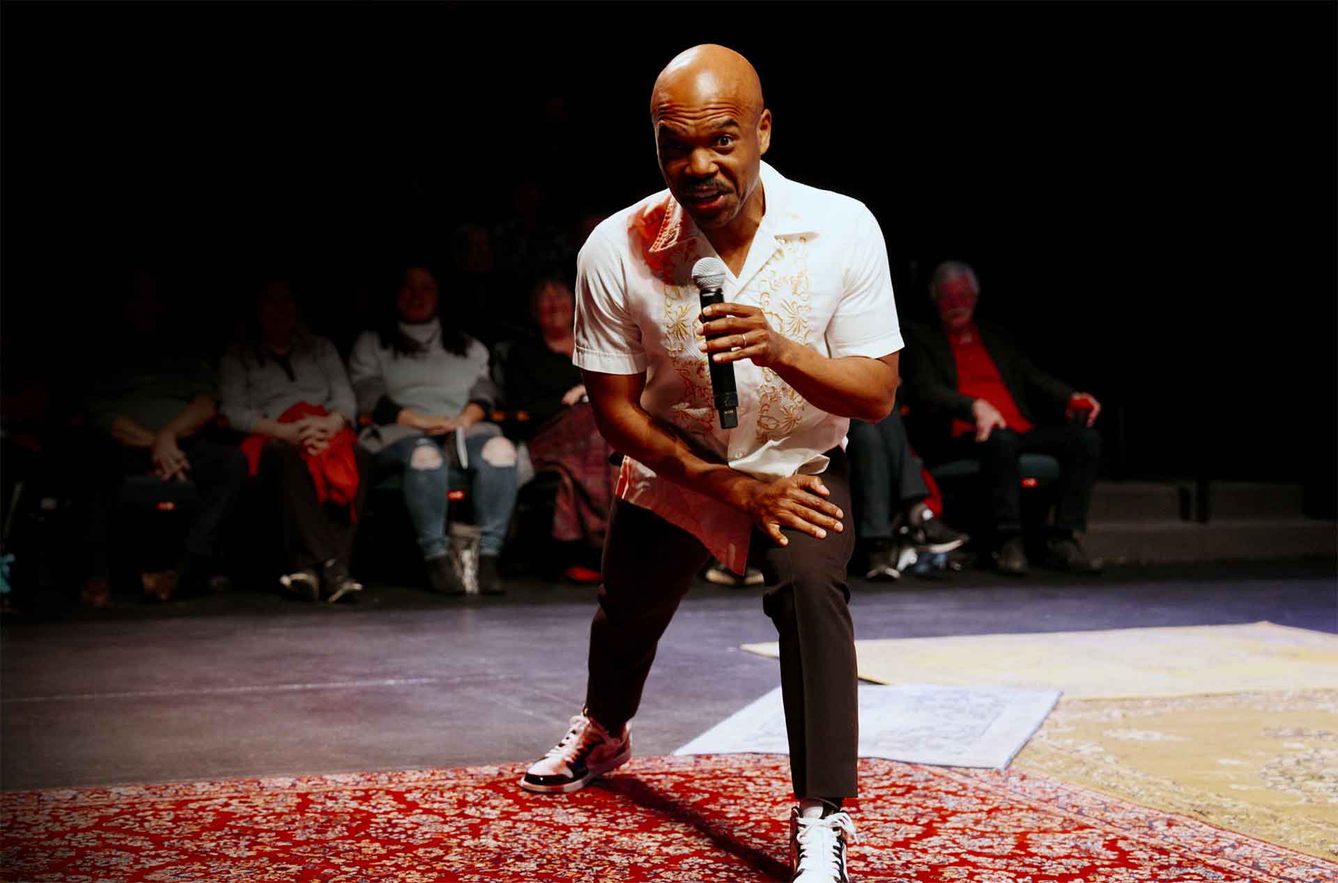 A performer holds a microphone while leaning toward the audience on a dimly lit stage.
