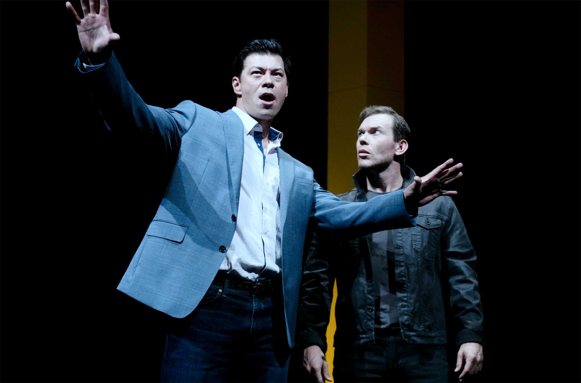 Two performers on a dark stage under spotlights, with one raising both arms while the other stands nearby looking upward.