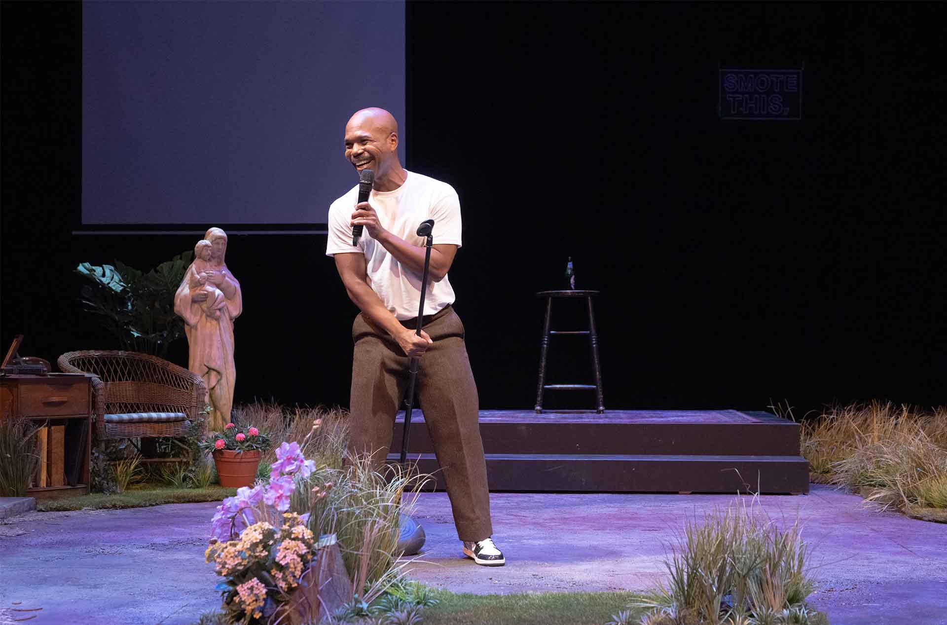 Performer smiles into a microphone onstage with a statue, flowers, stool, and dark backdrop behind.