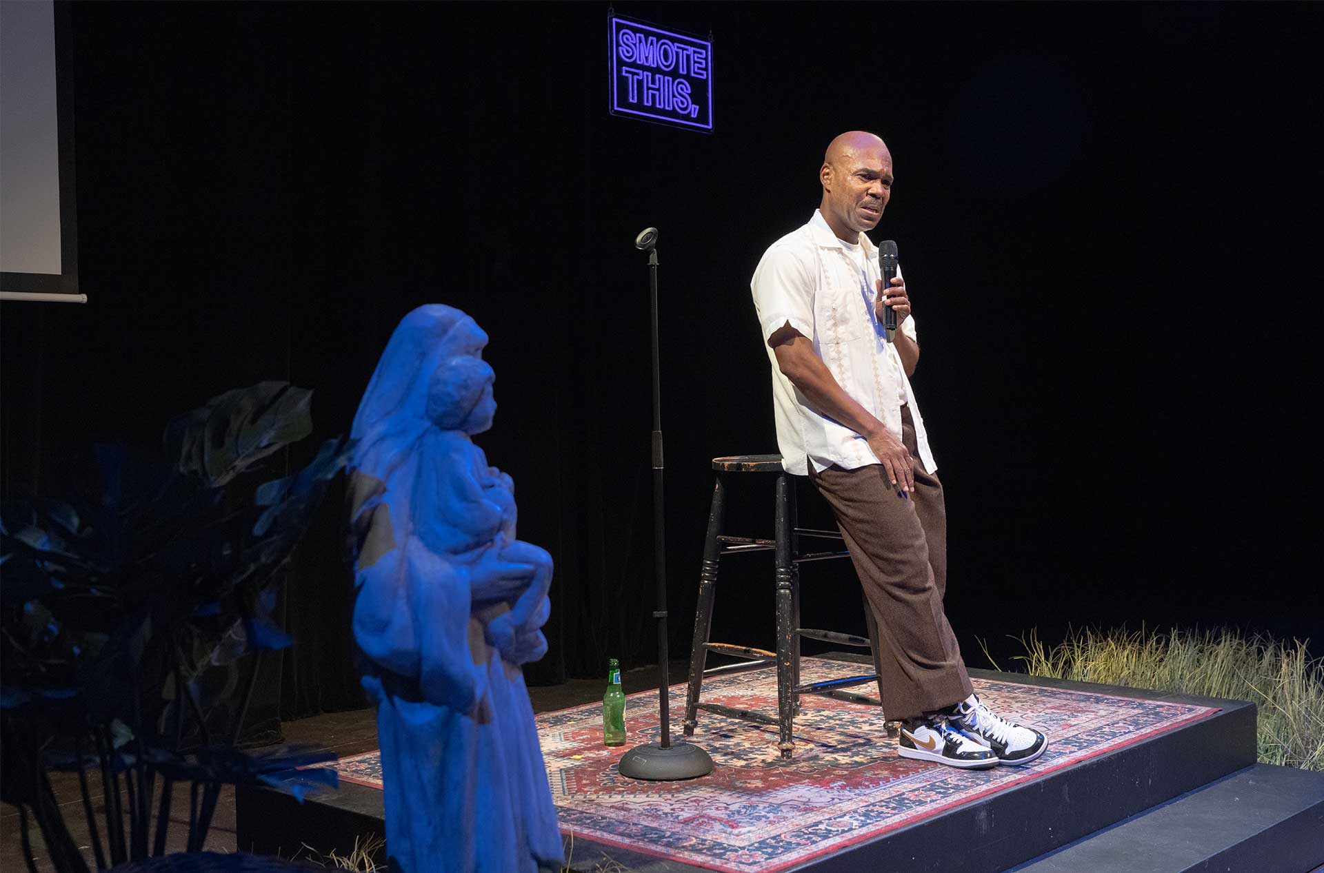 Performer stands beside a microphone and stool with a statue in the foreground and a glowing "SMOTE THIS." sign behind.