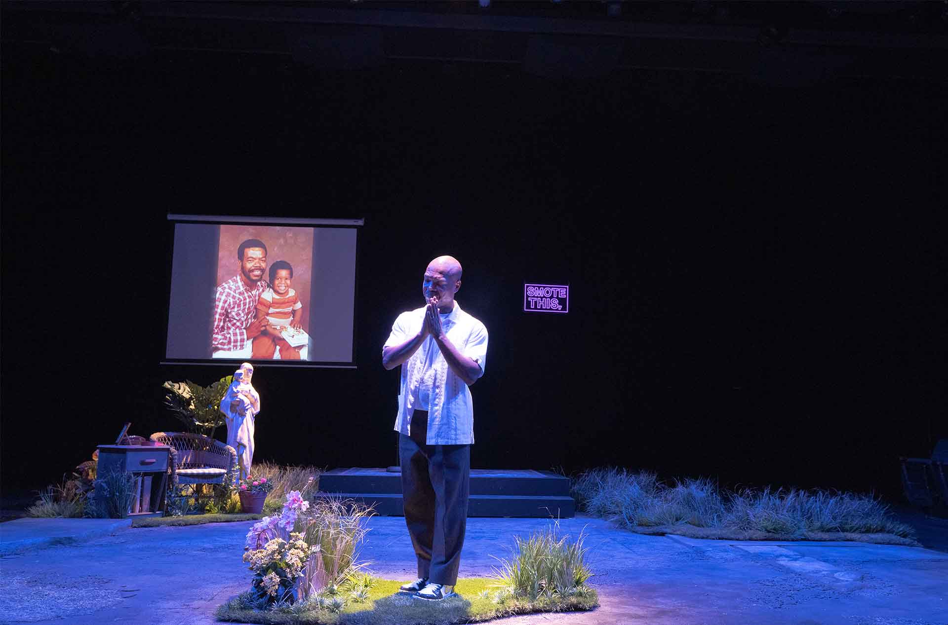 Performer stands with hands together beneath a projected family photo and the glowing words "SMOTE THIS."