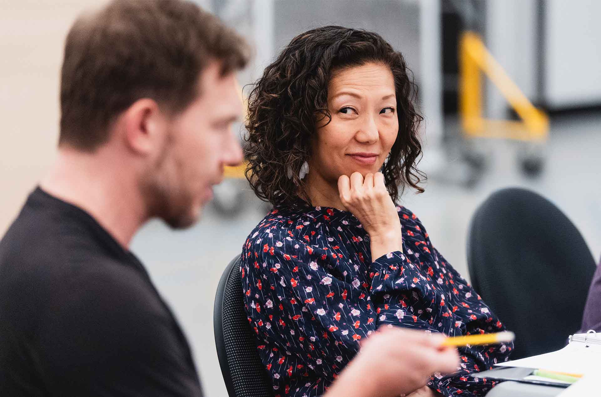 Two cast members sit at a rehearsal table; one smiles while listening.