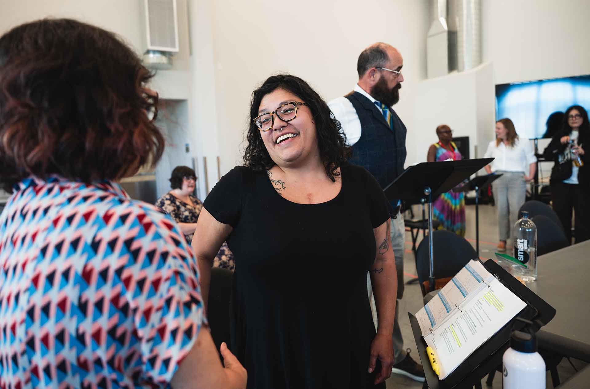 Cast  and creative team members laugh and talk during a rehearsal break, with music stands and binders nearby.