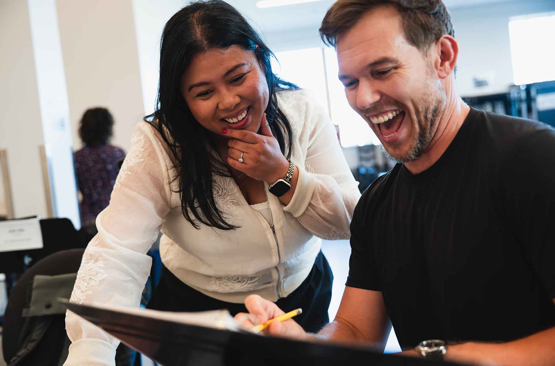 Two cast and creative team members smile while looking down at a rehearsal binder together.