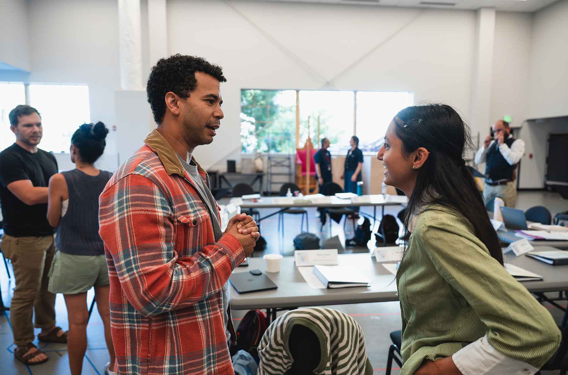 Two cast and creative team members talk in a rehearsal room while others gather in the background.