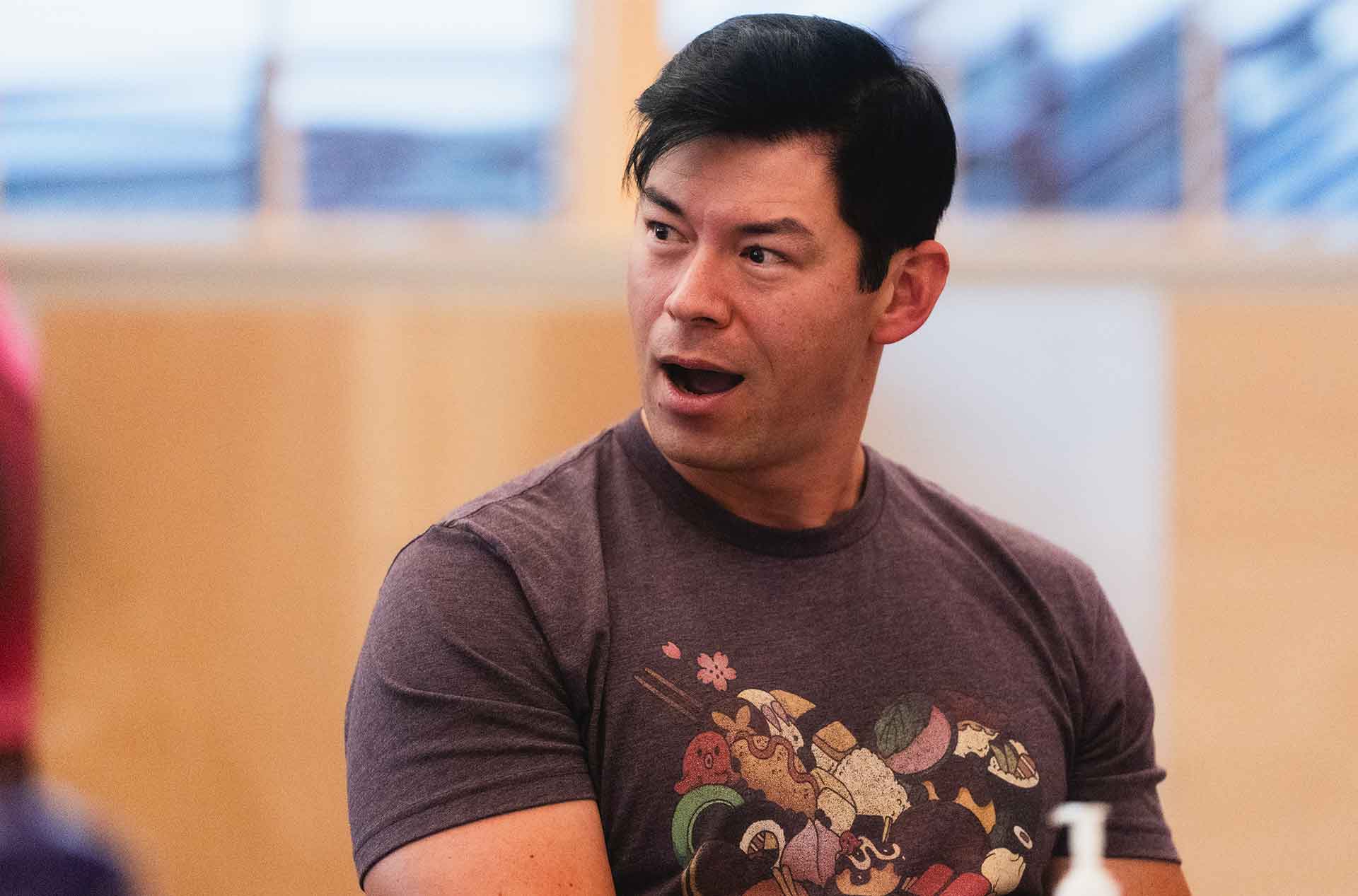 Cast member speaks during rehearsal, seated near a table with rehearsal materials.