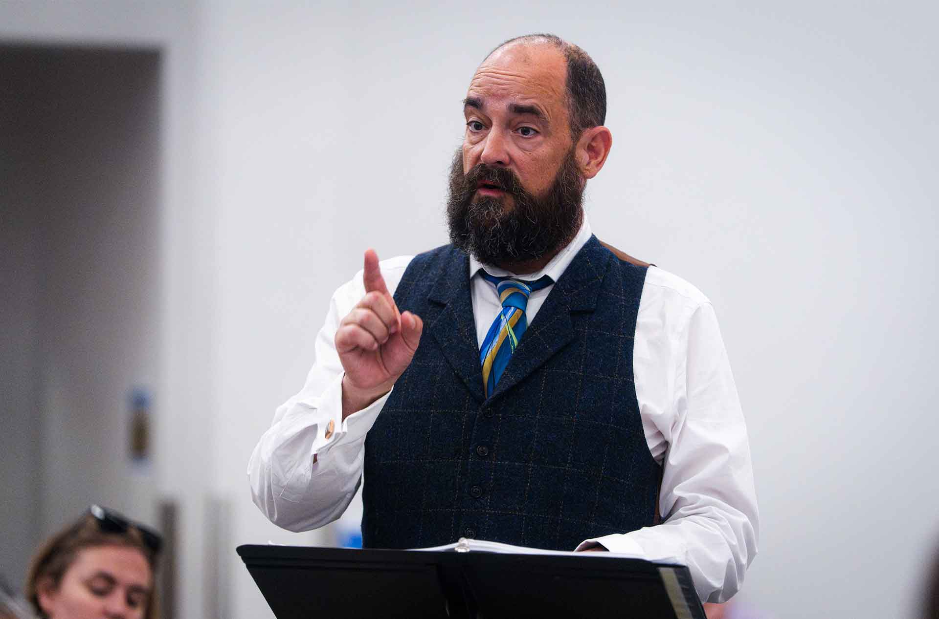 Person in a vest gestures while speaking from behind a music stand during rehearsal.