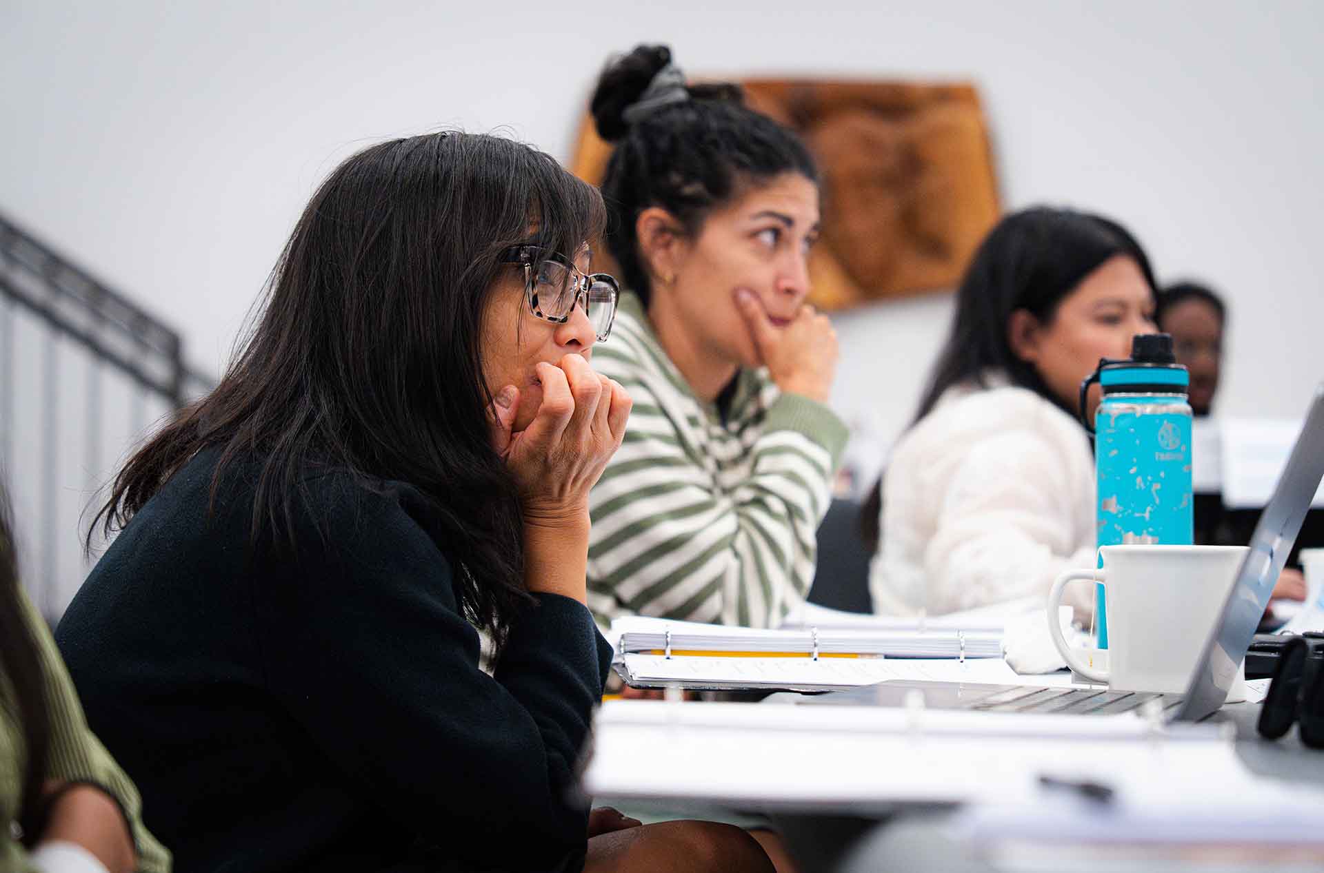 Several cast and creative team members sit at tables with binders and laptops, listening during rehearsal.