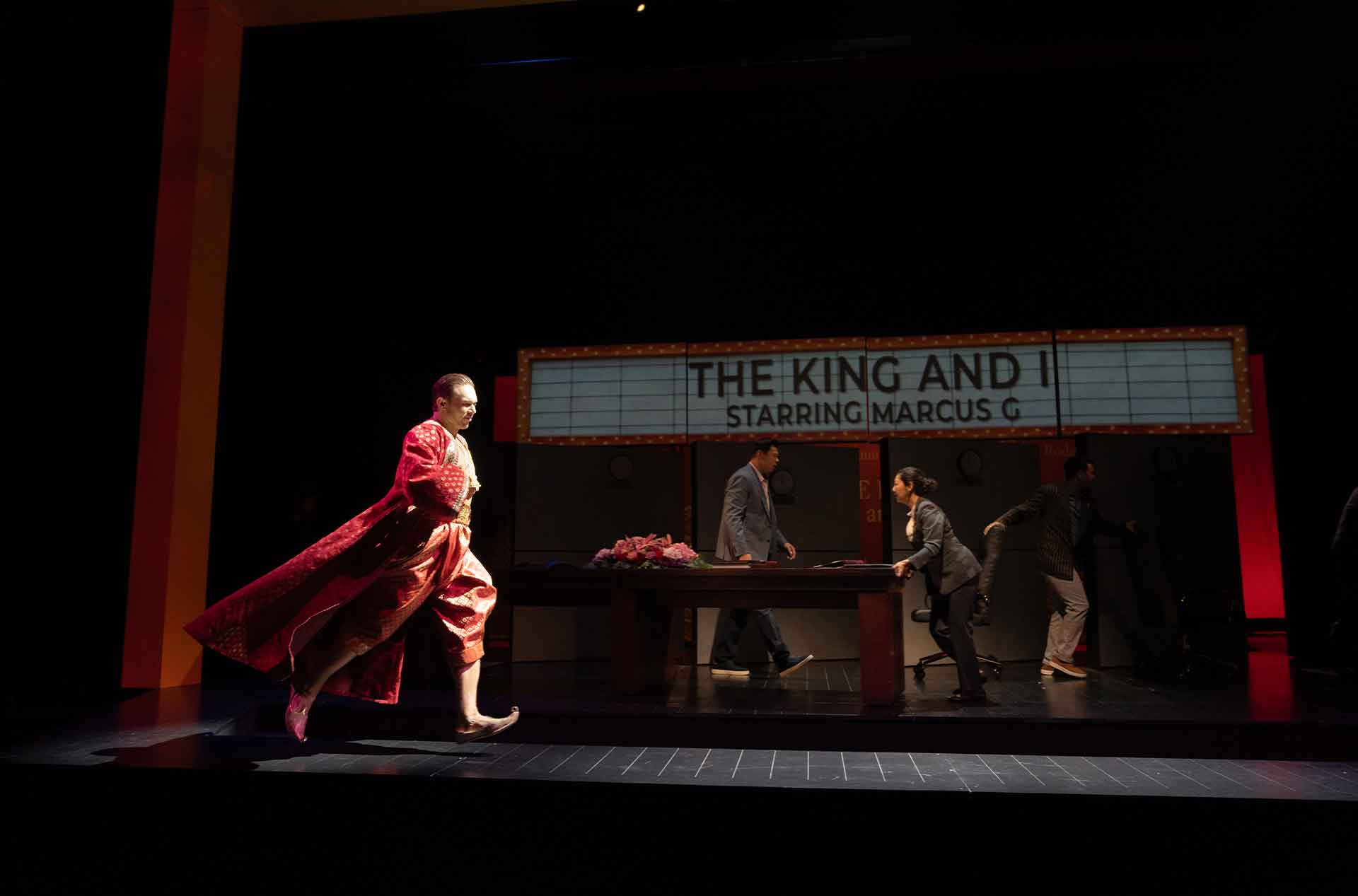 Alt text: A performer in an elaborate red and gold costume strides across the stage as several people move hurriedly around a table behind him. A theater marquee in the set reads "THE KING AND I" and "STARRING MARCUS G".