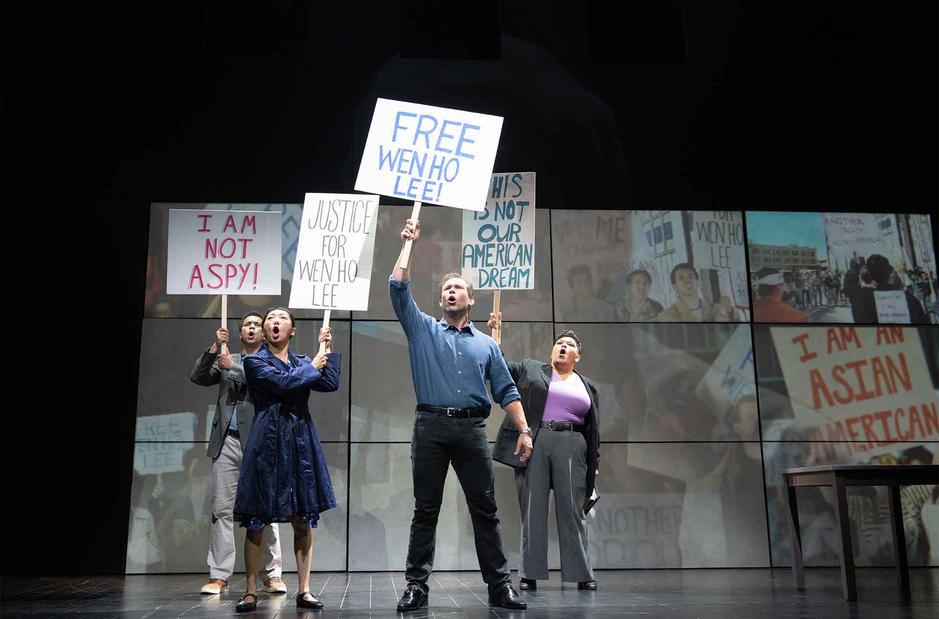 Four performers stand together on stage holding protest signs and shouting in front of projected demonstration scenes. The signs read "I AM NOT A SPY" "JUSTICE FOR WENHO LEE" "FREE WENHO LEE" and "THIS IS NOT OUR AMERICAN DREAM".