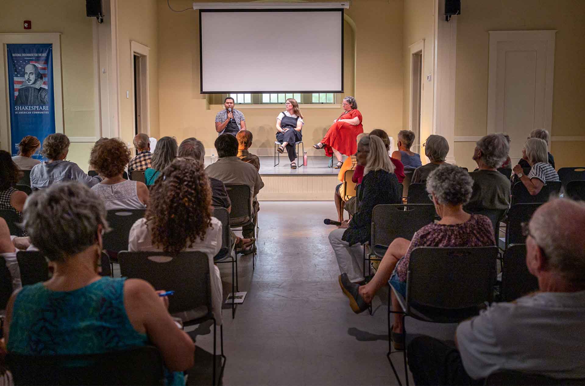 A group of people watches three speakers on a stage during a community panel discussion in a small event space. One speaker holds a microphone while answering questions.