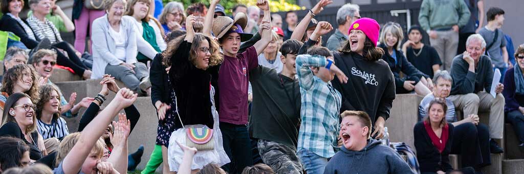 Large group of people sitting on outdoor steps cheering with raised arms during a lively community event. Several children stand in the foreground celebrating while others smile and clap in the background.