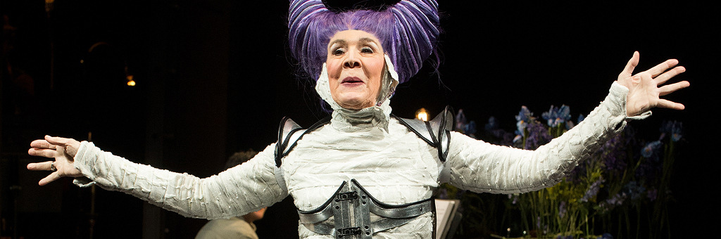 Performer in an elaborate stage costume with a sculptural purple headdress, standing with arms outstretched under theatrical lighting.