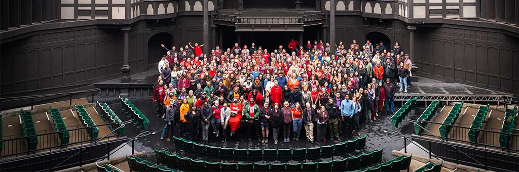 A large group of people gathered on the stage of an outdoor Tudor-style theater, with dramatic cloudy skies and green stadium seating in the foreground.